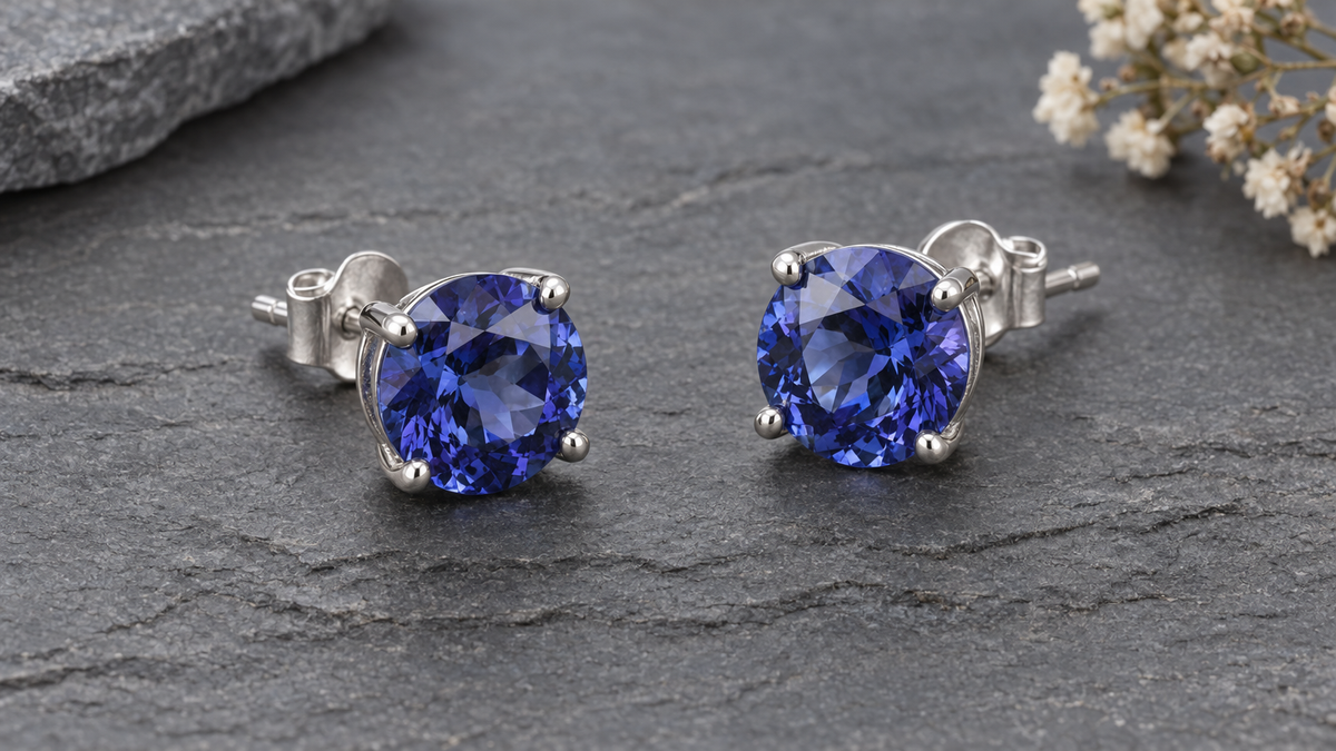 Illustrative still life of silver-tone stud earrings with round blue stones on dark grey slate. This is illustrative only, not a verified depiction of specific Awareness Avenue products, tanzanite, or materials.