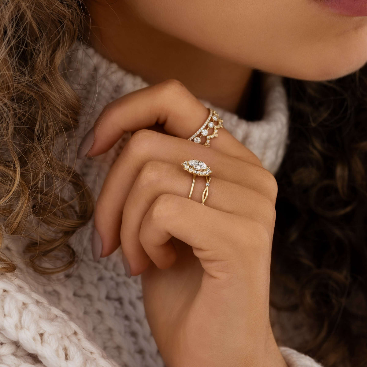 Close-up of a hand at a jaw wearing gold-toned stacked rings, including one with an oval halo stone.