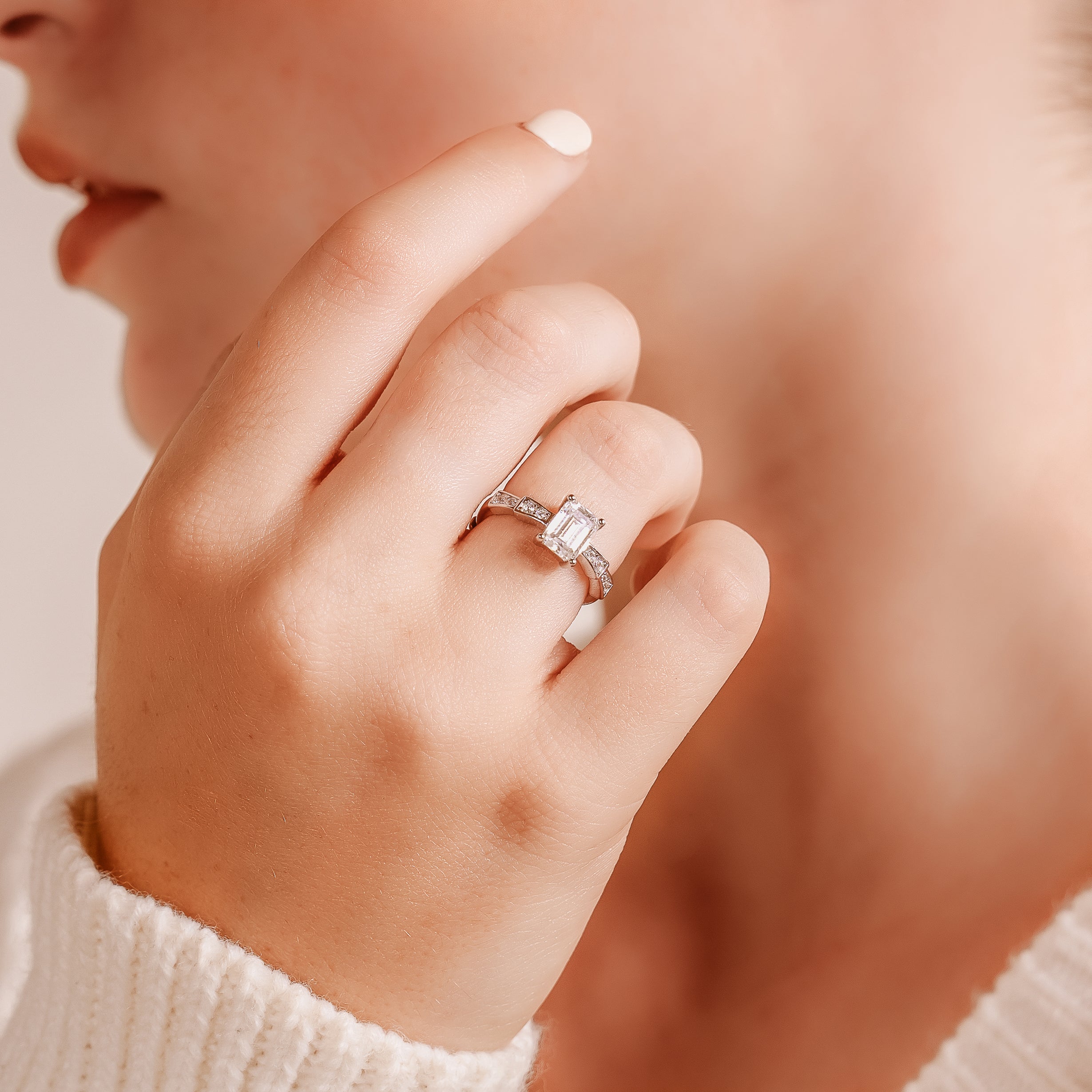 Close-up of a hand near a face, fingertip with cream and a silver-toned ring with emerald-cut center stone.