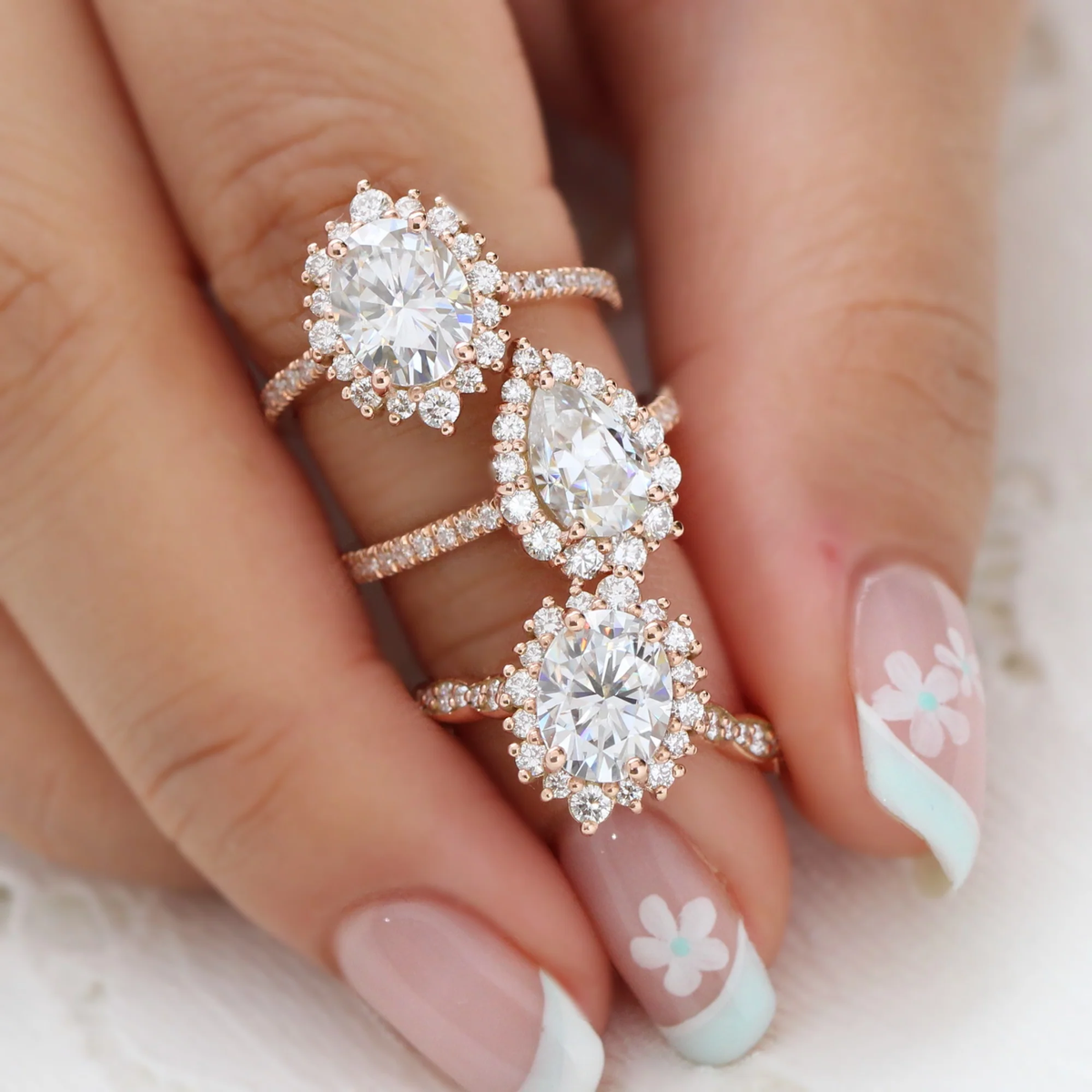 Three gold-toned halo rings with clear oval and pear gems stacked on a manicured hand.