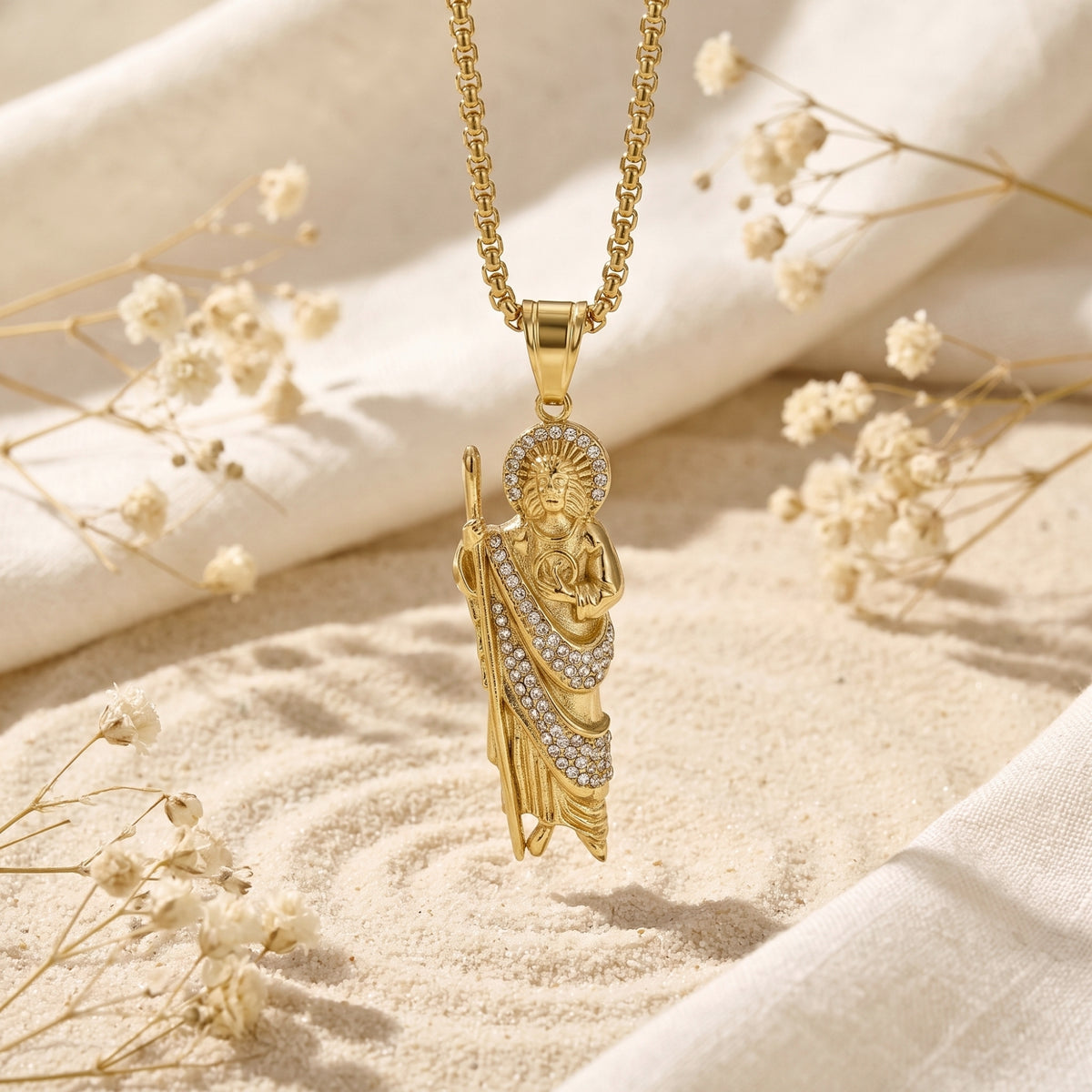 Gold-tone St. Jude pendant necklace suspended over sand and dried white flowers.