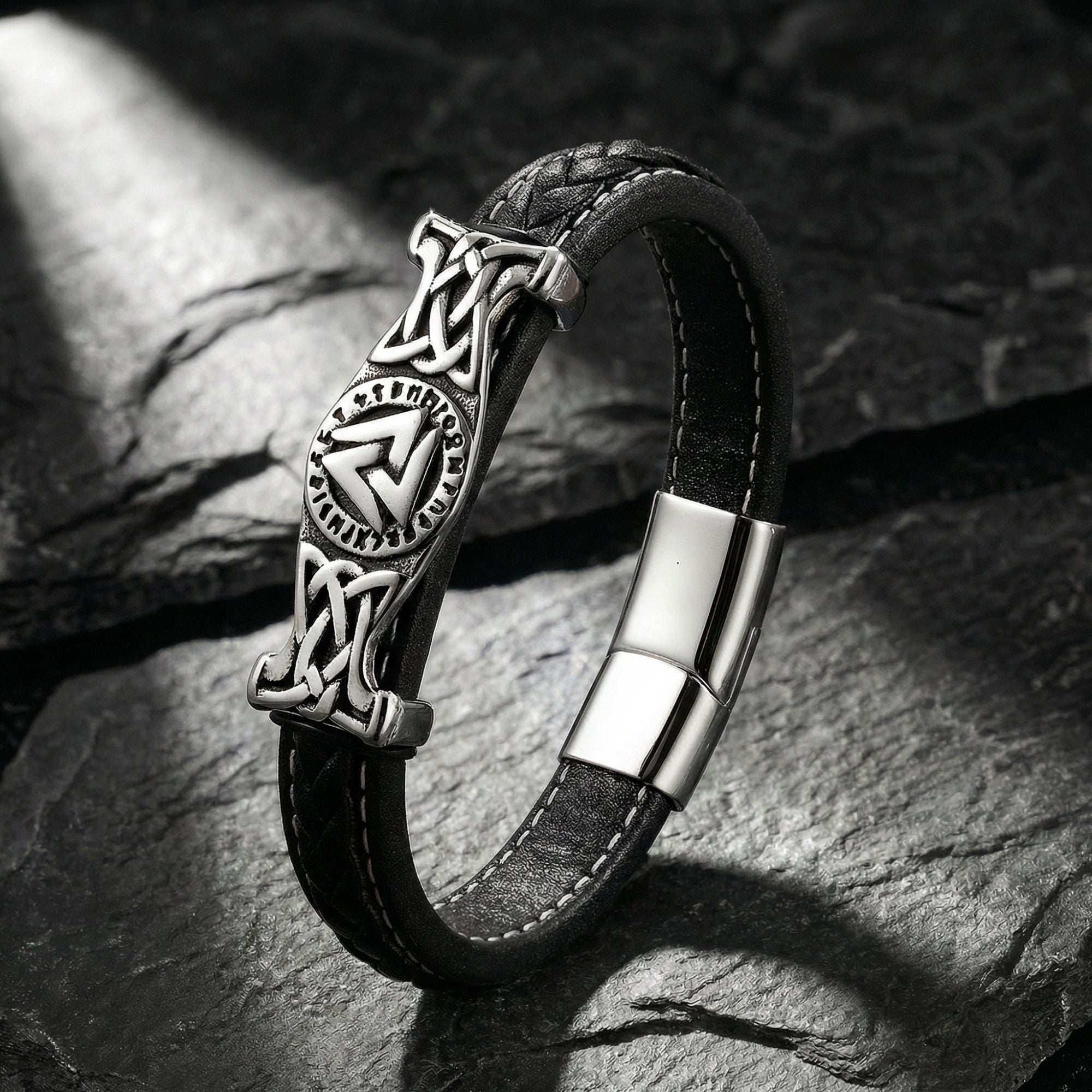 Black braided leather Runeknot bracelet with stainless steel Valknut centerpiece on a dark stone background.
