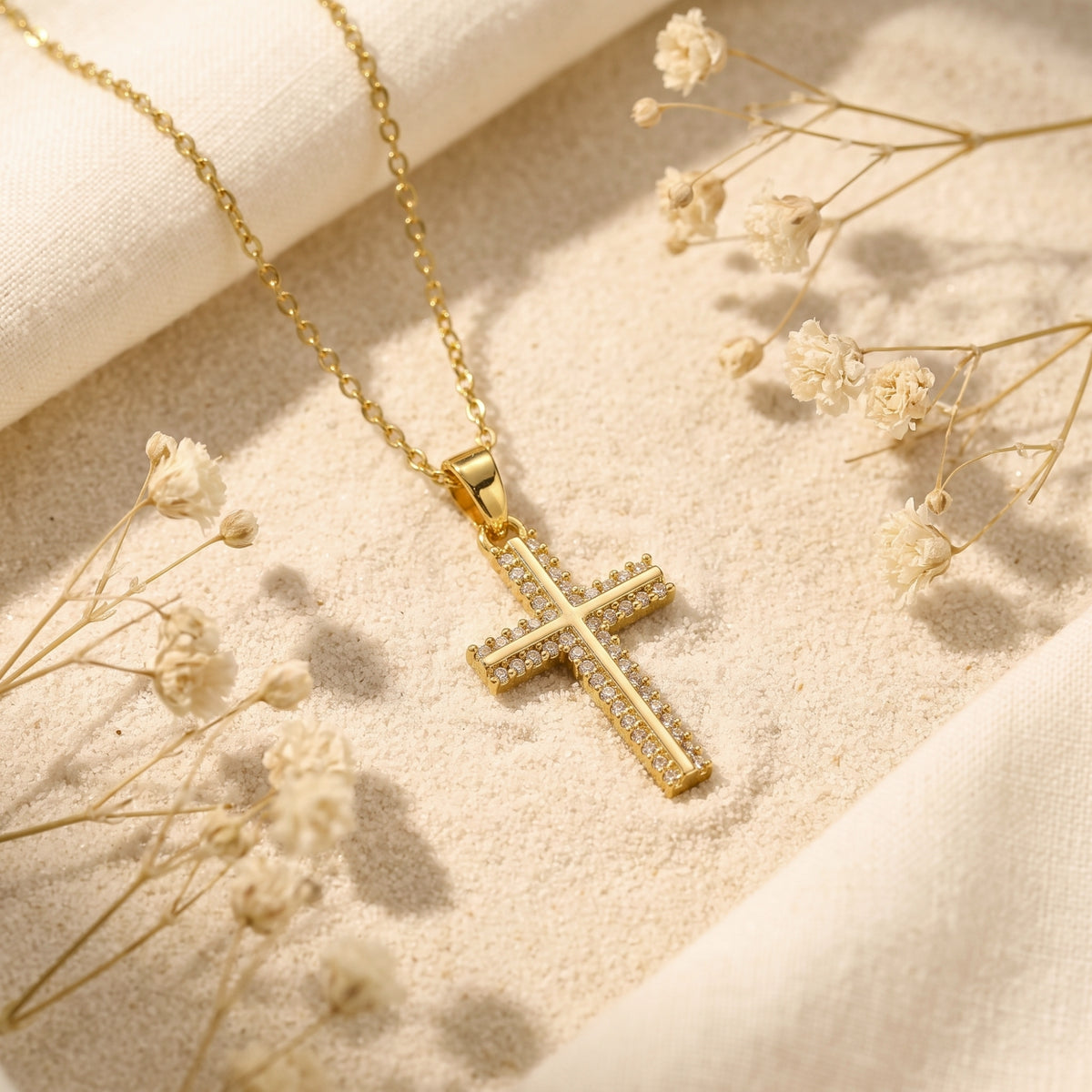 Gold-tone pavé cross necklace with clear cubic zirconia on sand among dried flowers.