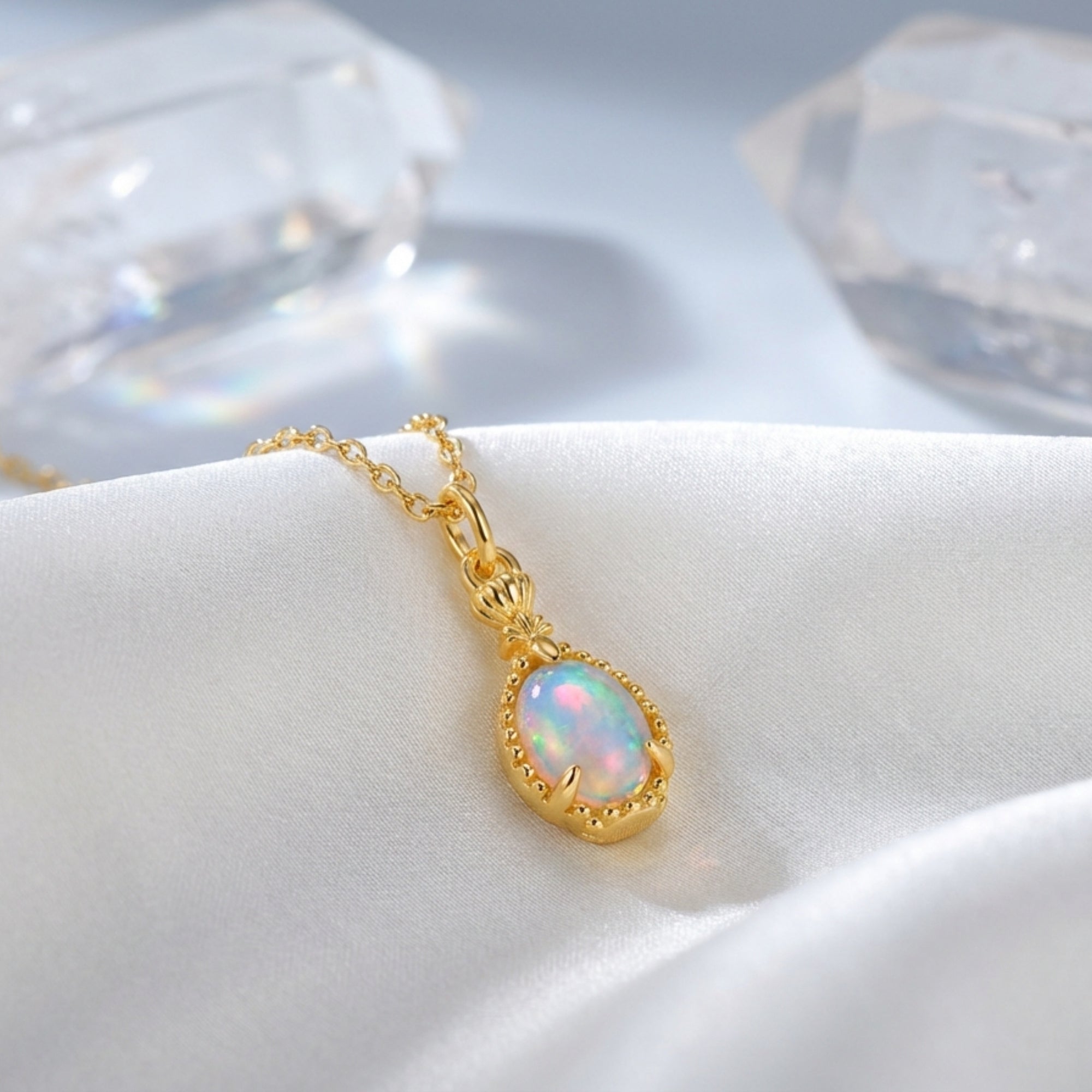 Gold-tone pear cut natural opal pendant necklace resting on white silk fabric with decorative crystals.