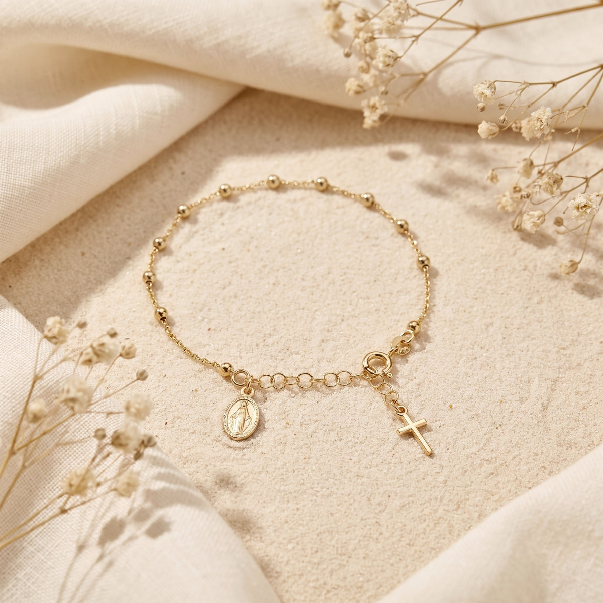 Gold beaded charm bracelet with medal and cross, laid flat on sand beside linen fabric.