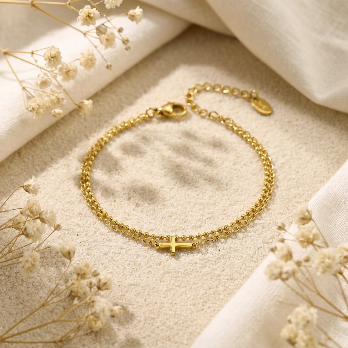 Gold-finish double-chain cross bracelet arranged in a circle on sand with dried flowers and linen.