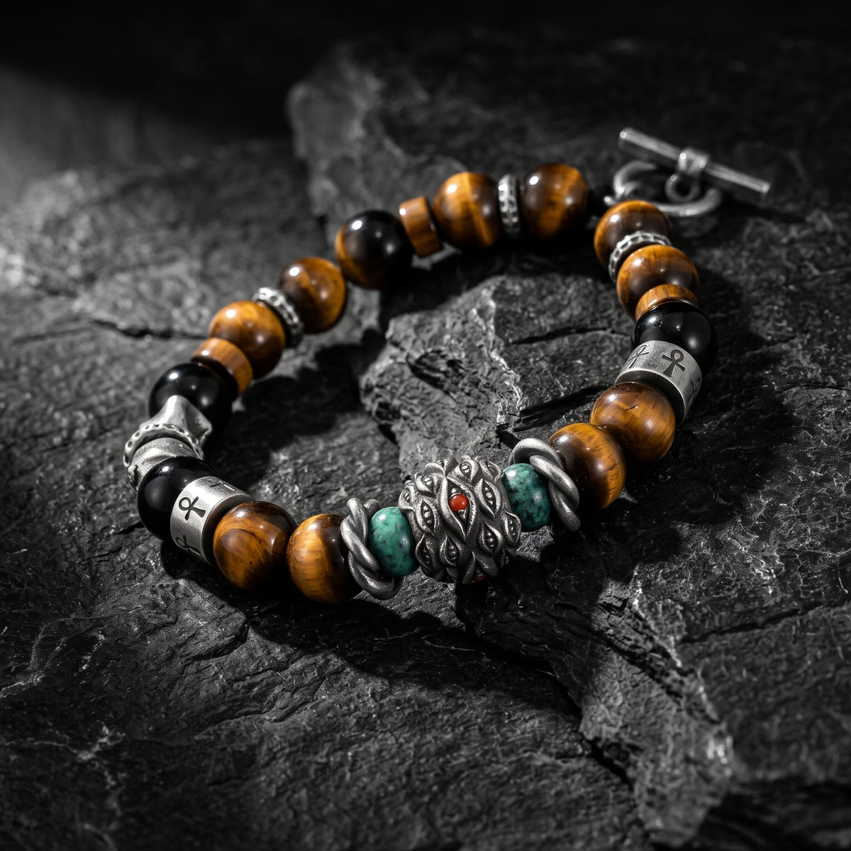 Angled view of a silver beaded bracelet with tiger's eye, ankh spacers, and an eye charm on dark slate.