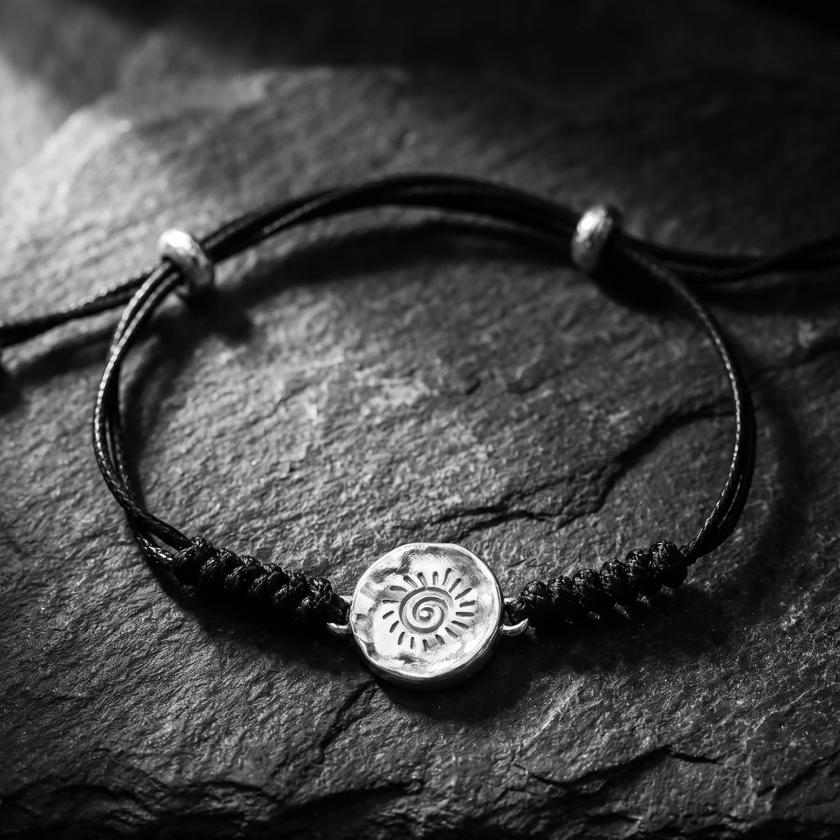 Sol cord bracelet with a round silver sun medallion displayed on dark slate.