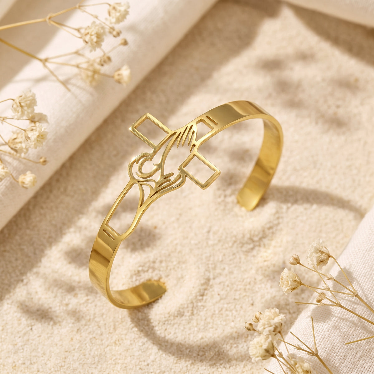 Gold-tone praying hands and cross cuff bracelet resting on sand beside dried white flowers.