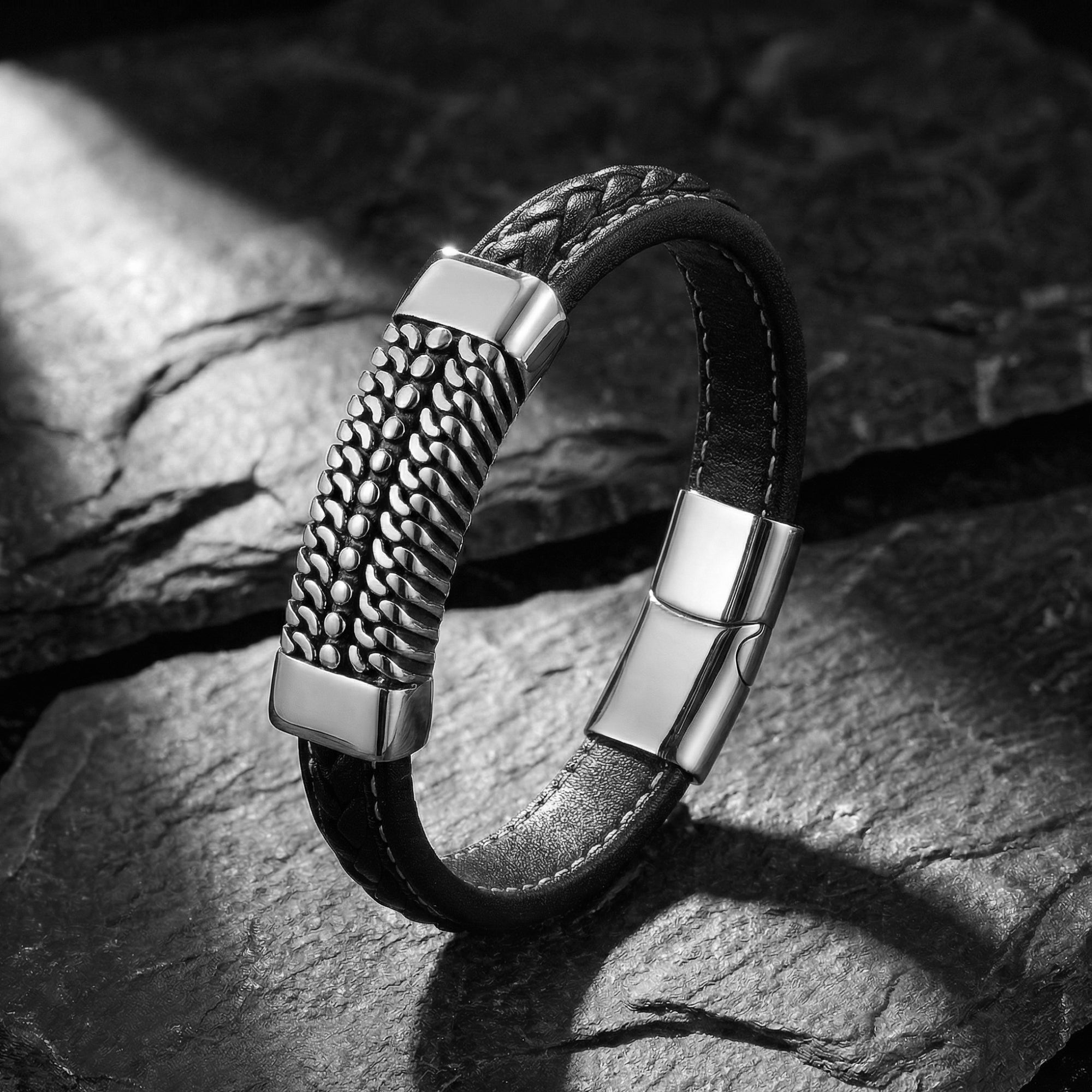 Black braided leather Kinetic Bracelet with a textured stainless steel center plate on a dark stone background.
