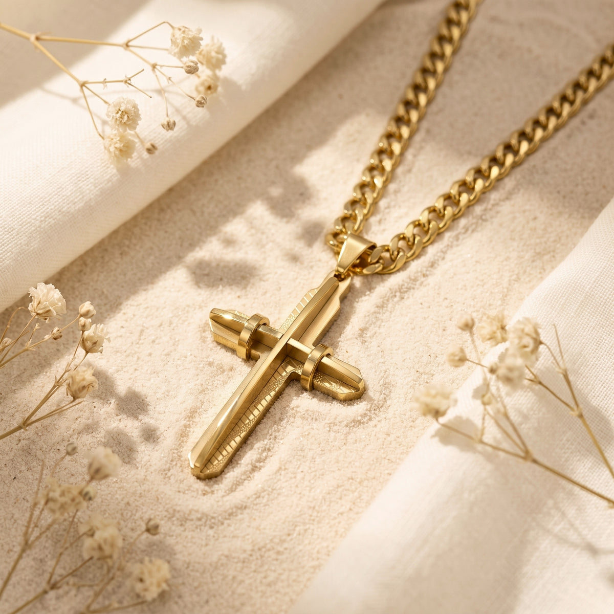 Gold-toned layered cross pendant with curb chain lying on pale sand among dried white flowers.