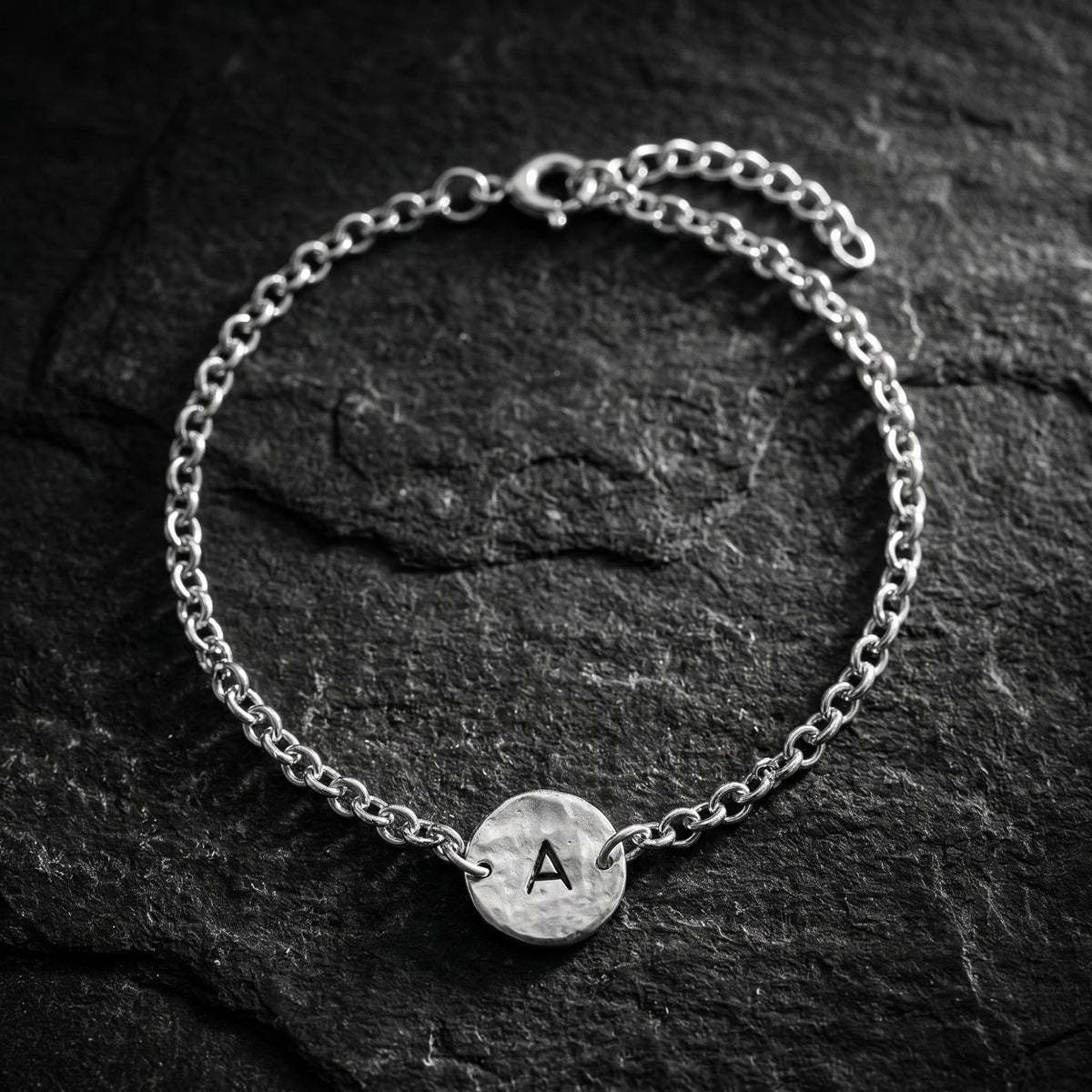 Top-down view of a silver chain bracelet with a hammered round disc stamped with initial A on dark slate.