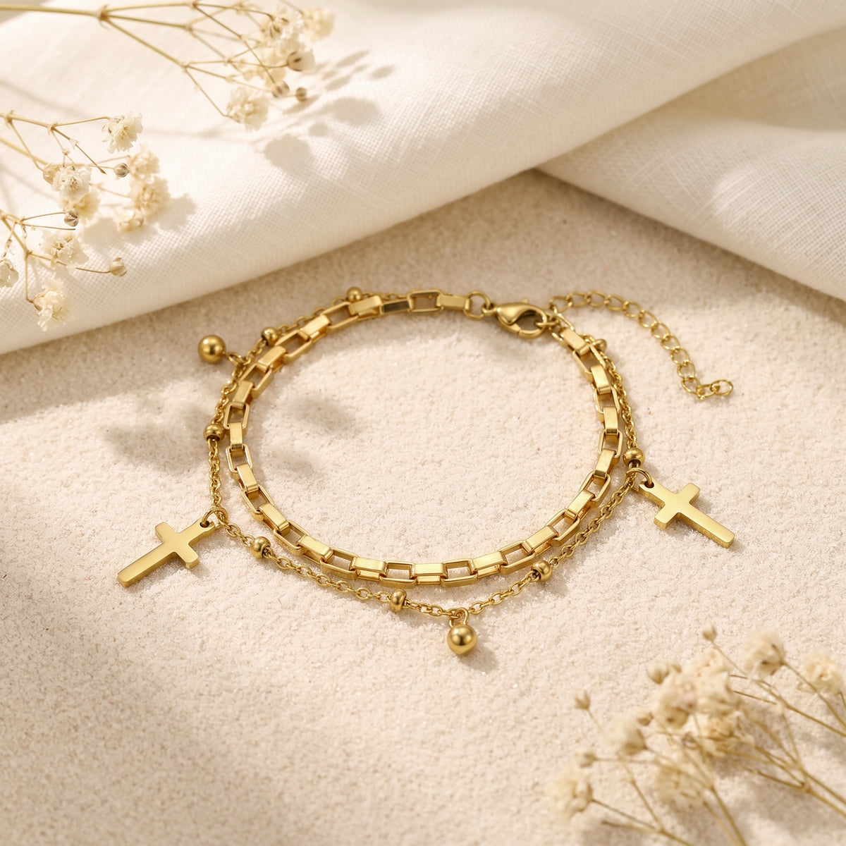 Gold-tone double-layered cross bracelet laid flat on a textured surface beside white cloth and dried flowers.
