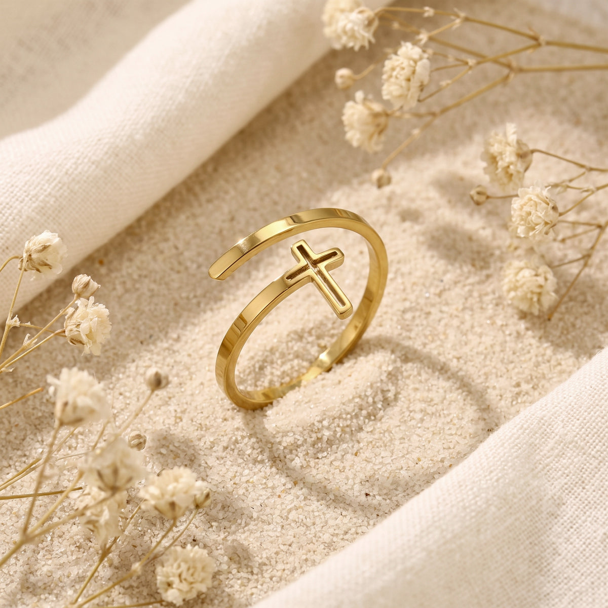 Gold-finish open-wrap cross ring displayed upright on sand among dried white flowers.