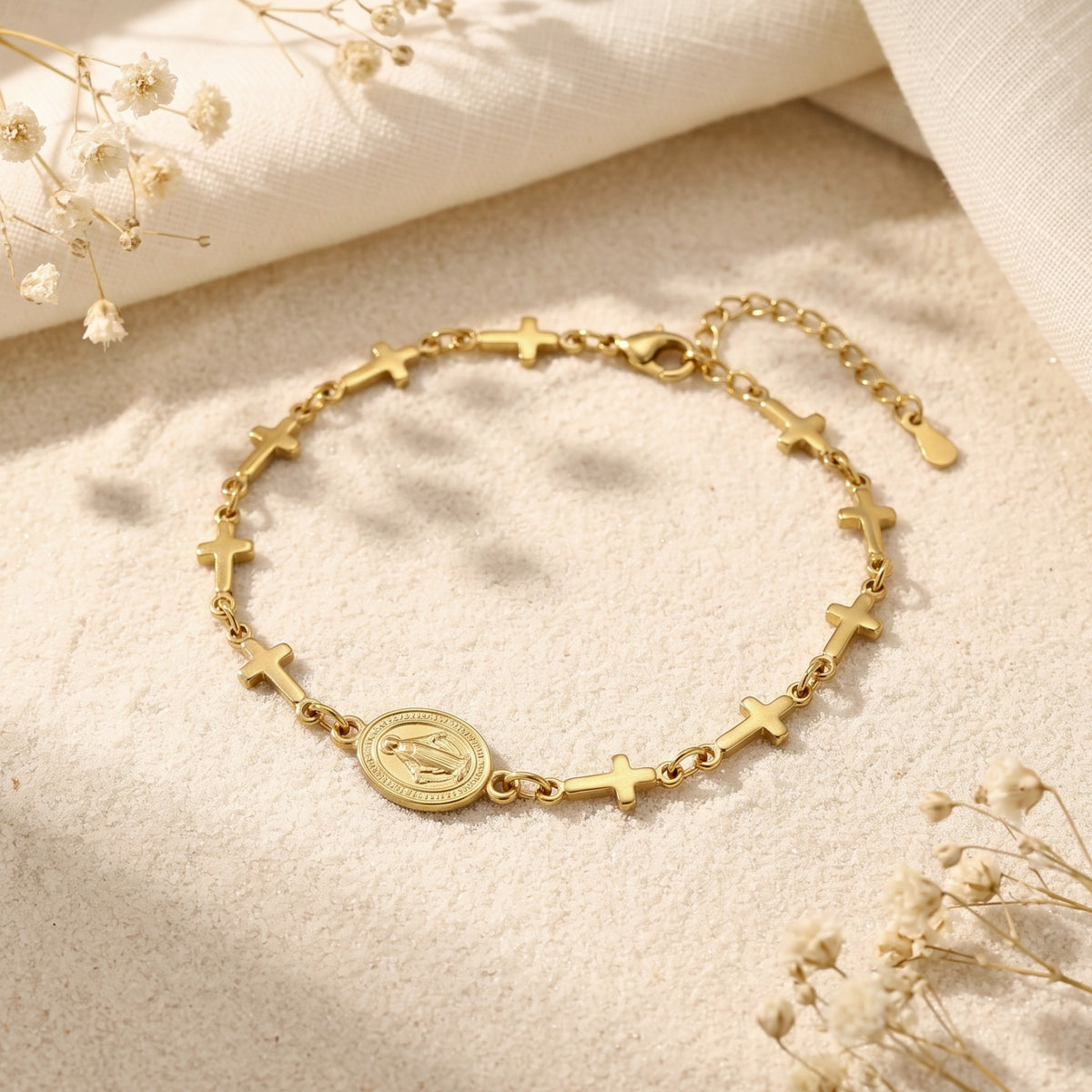Gold-tone cross and oval medal station bracelet arranged flat on a sandy surface with white linen.
