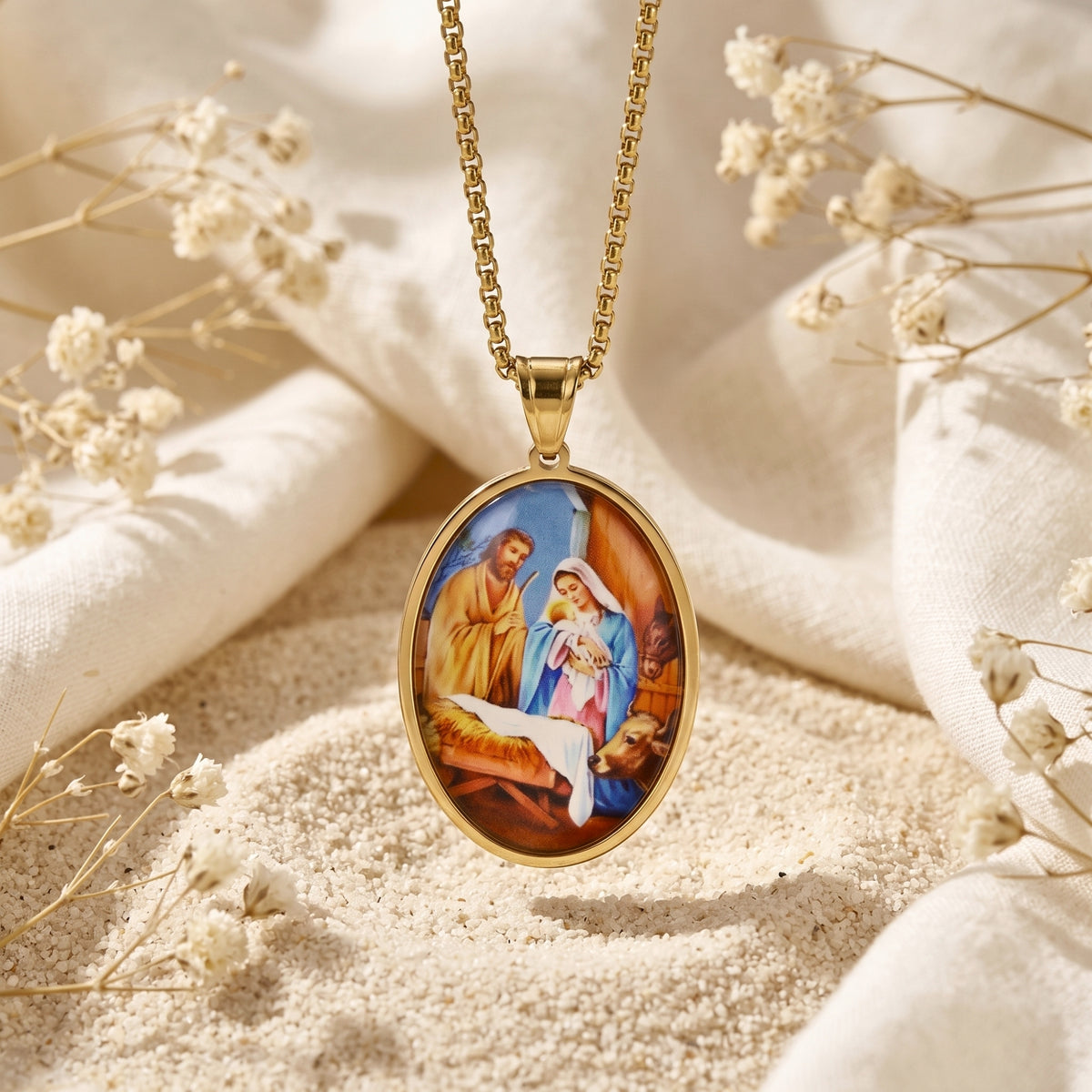 Gold-tone oval Nativity pendant necklace hanging vertically over pale sand, white fabric, and dried flowers.