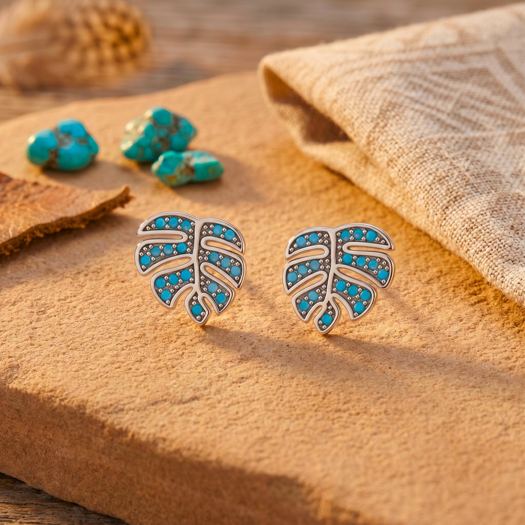 Silver monstera leaf earrings with turquoise cabochons styled on a desert stone with raw turquoise.