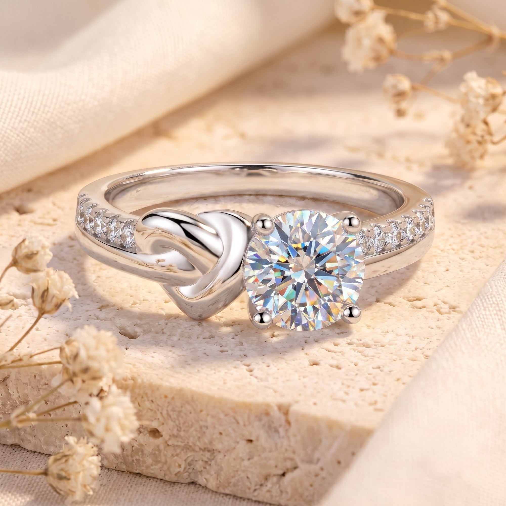 Silver love knot promise ring with round-cut cubic zirconia on a stone surface with dried flowers.