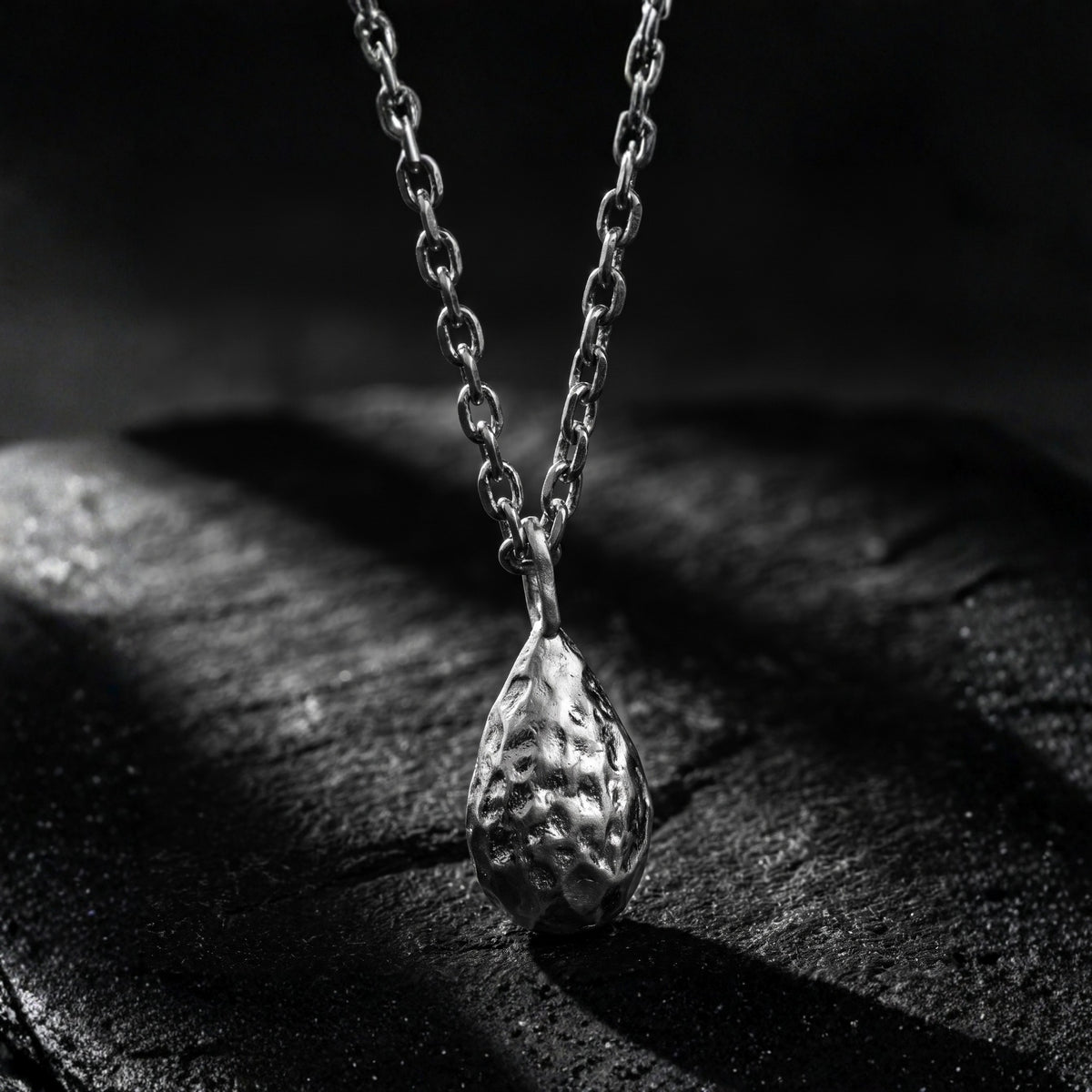 Hammered silver teardrop pendant attached to a link chain on a dark textured surface.