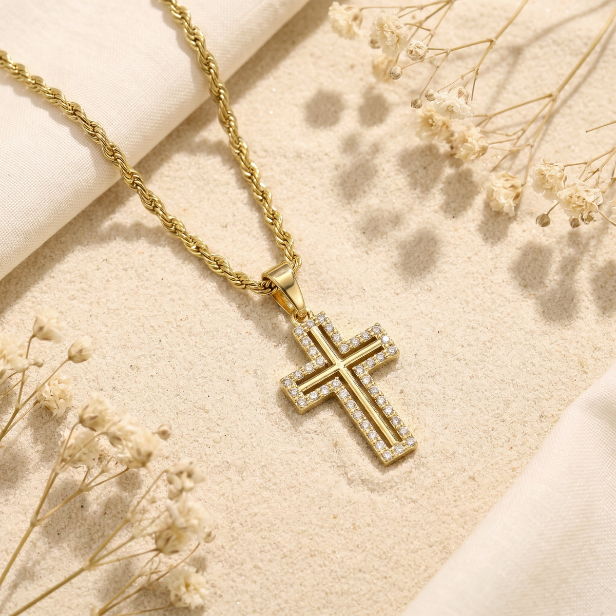 Gold-tone rope chain and cross pendant with stone-set border lying on sand with dried flowers.