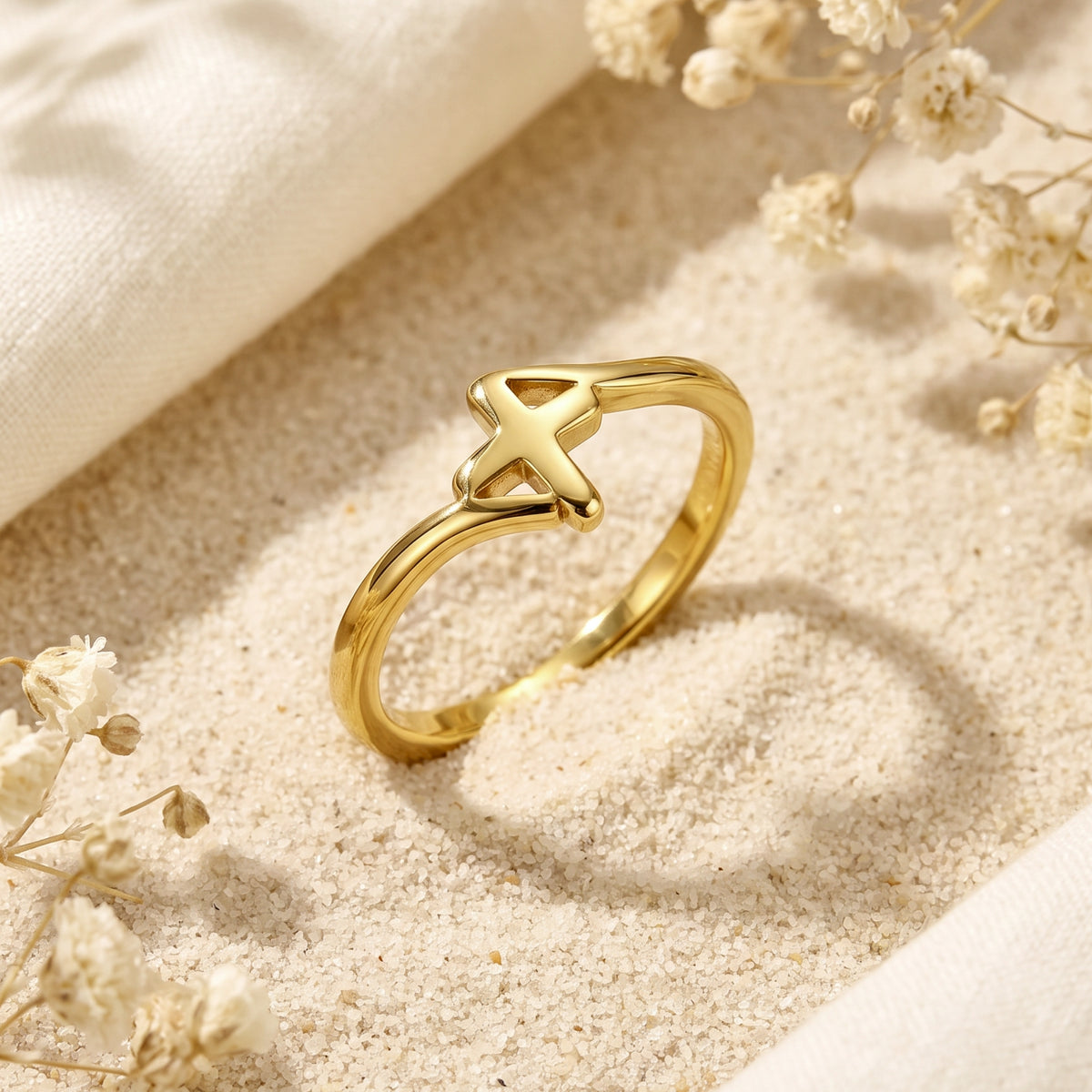 Angled view of a gold-tone openwork cross ring resting in fine sand with dried flowers.