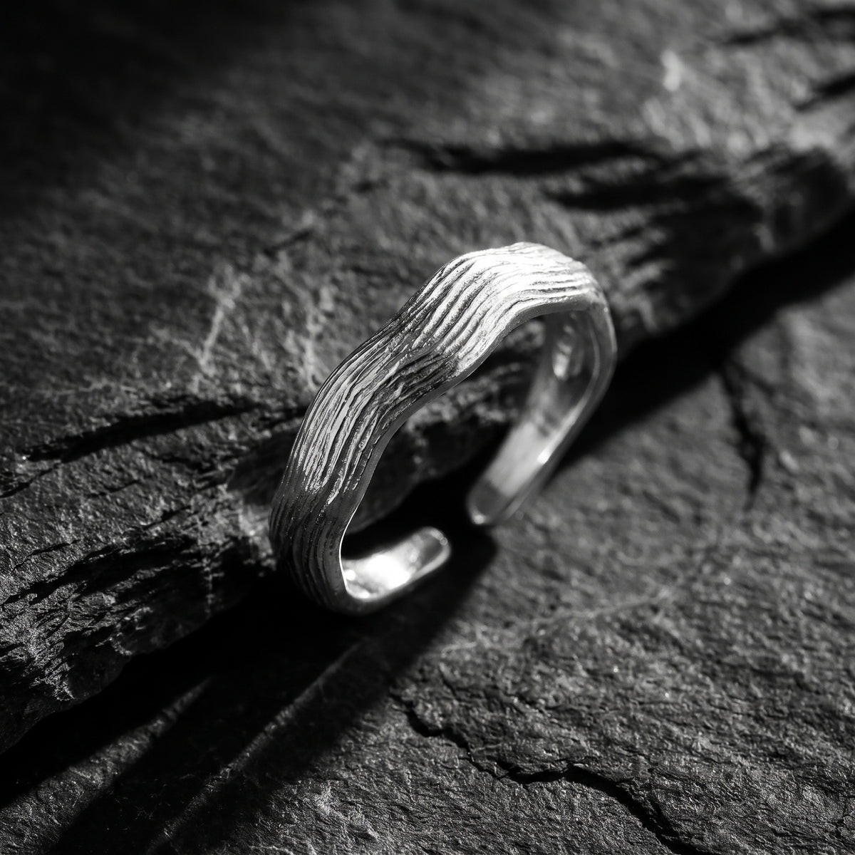 Angled close-up of a silver open-band ring with carved wave texture resting on dark slate.