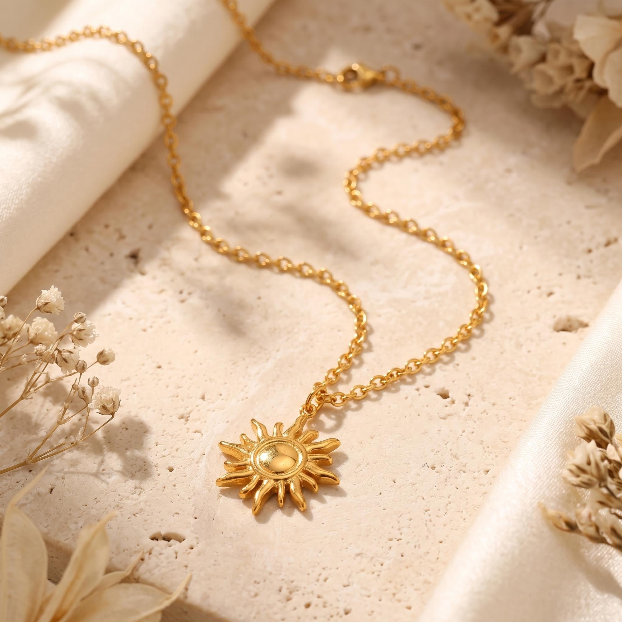 Gold-tone sun pendant necklace displayed on a textured beige surface beside cream fabric and dried flowers.