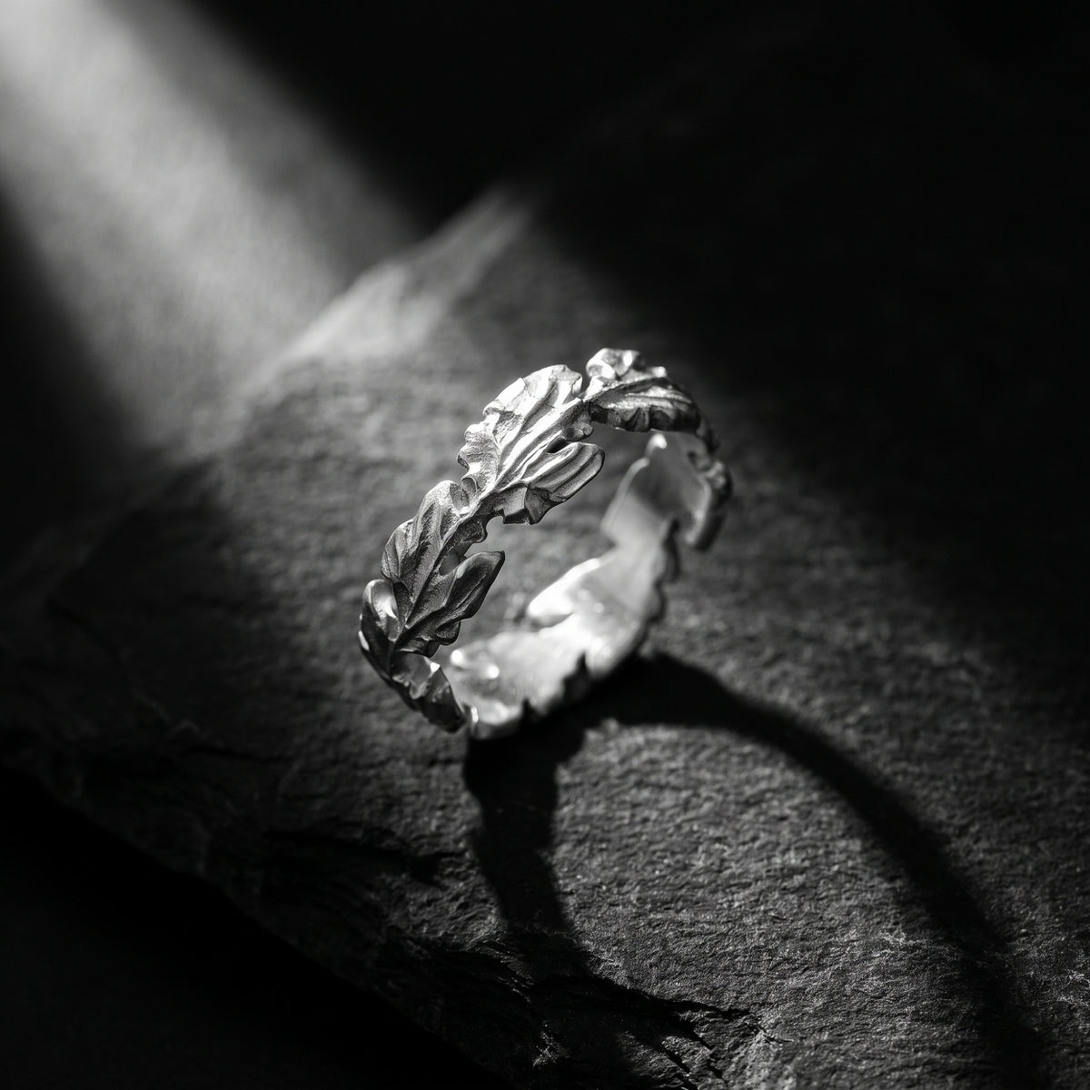 Silver oak leaf ring tilted upright on a dark textured surface under a strong spotlight.