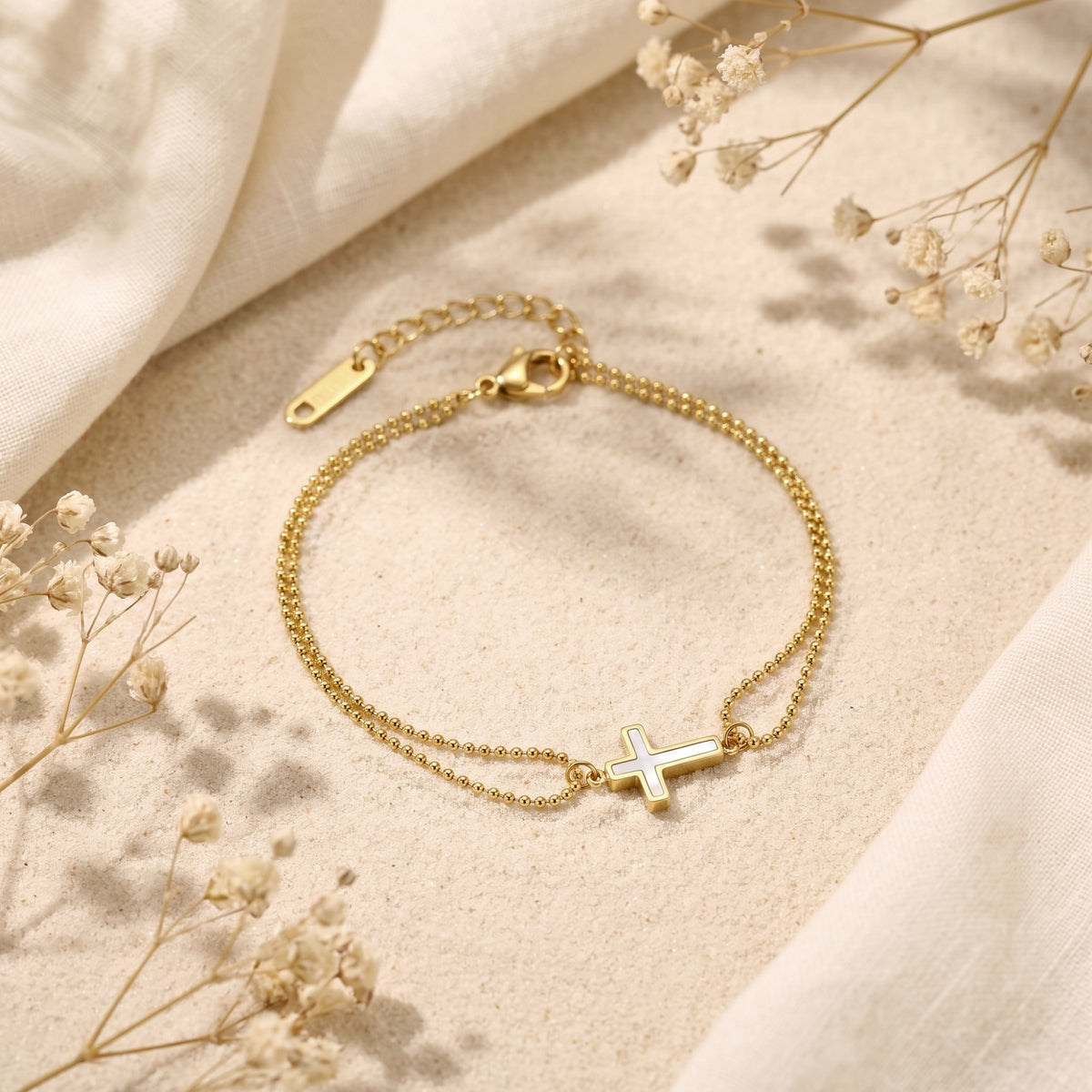Gold double-bead bracelet with a mother-of-pearl cross charm displayed on sand-colored fabric with dried flowers.