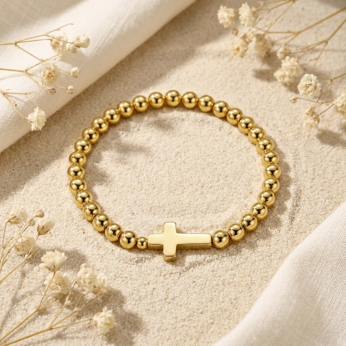 Overhead view of a gold-tone beaded bracelet with a smooth horizontal cross, lying flat on sand among dried flowers.