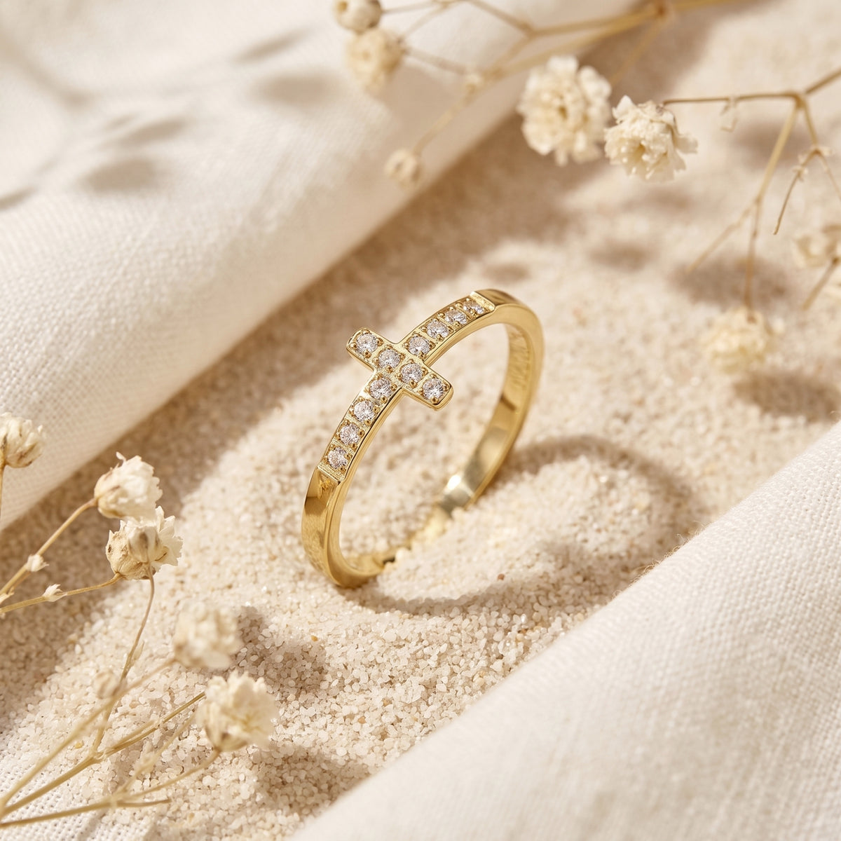 Angled close-up of a gold-tone cross ring with clear crystals resting on sand beside dried white flowers.