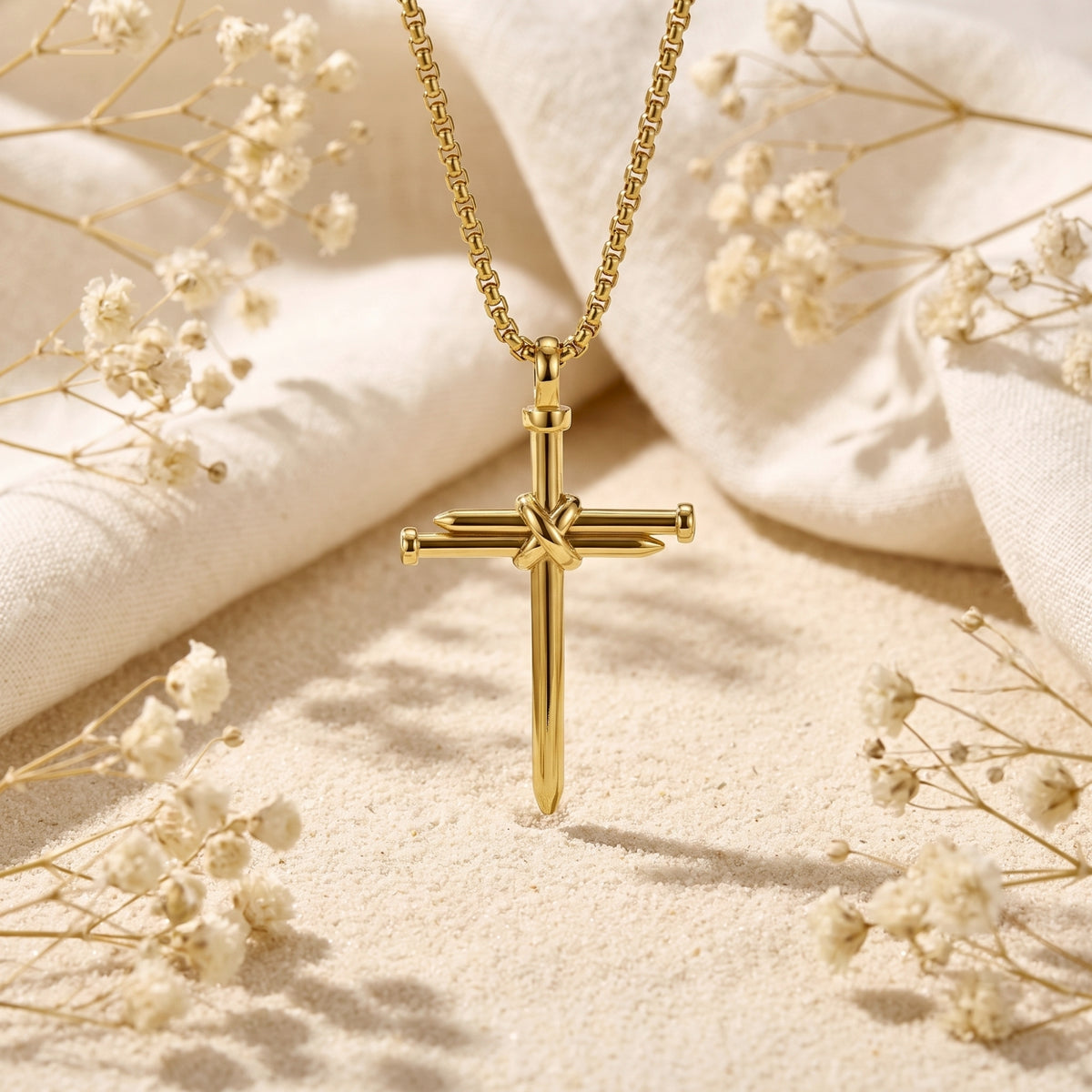 Gold-tone bound nail cross pendant hanging above sand, framed by white fabric and dried flowers.
