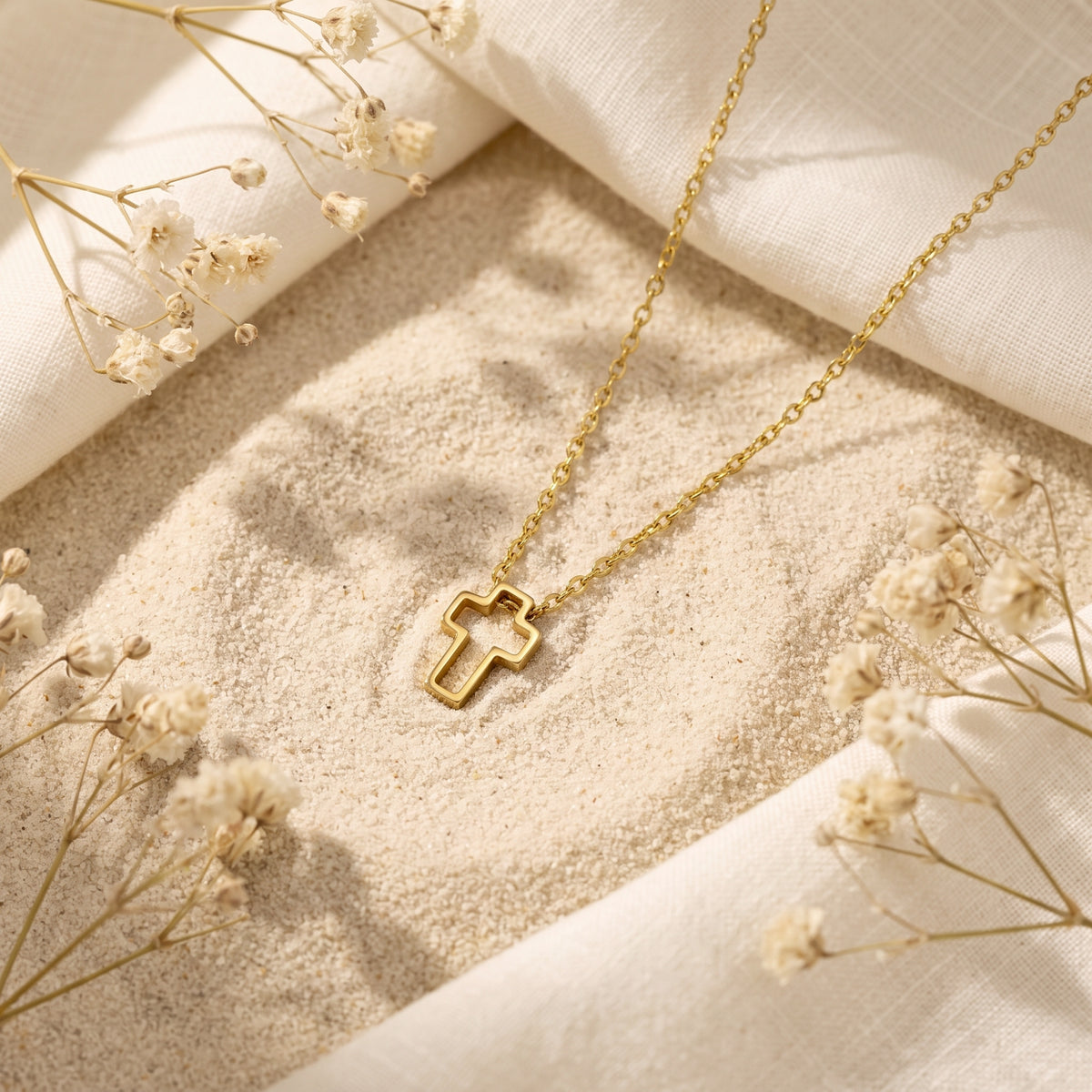 Gold-tone openwork cross necklace lying on sand with white fabric and dried flowers.