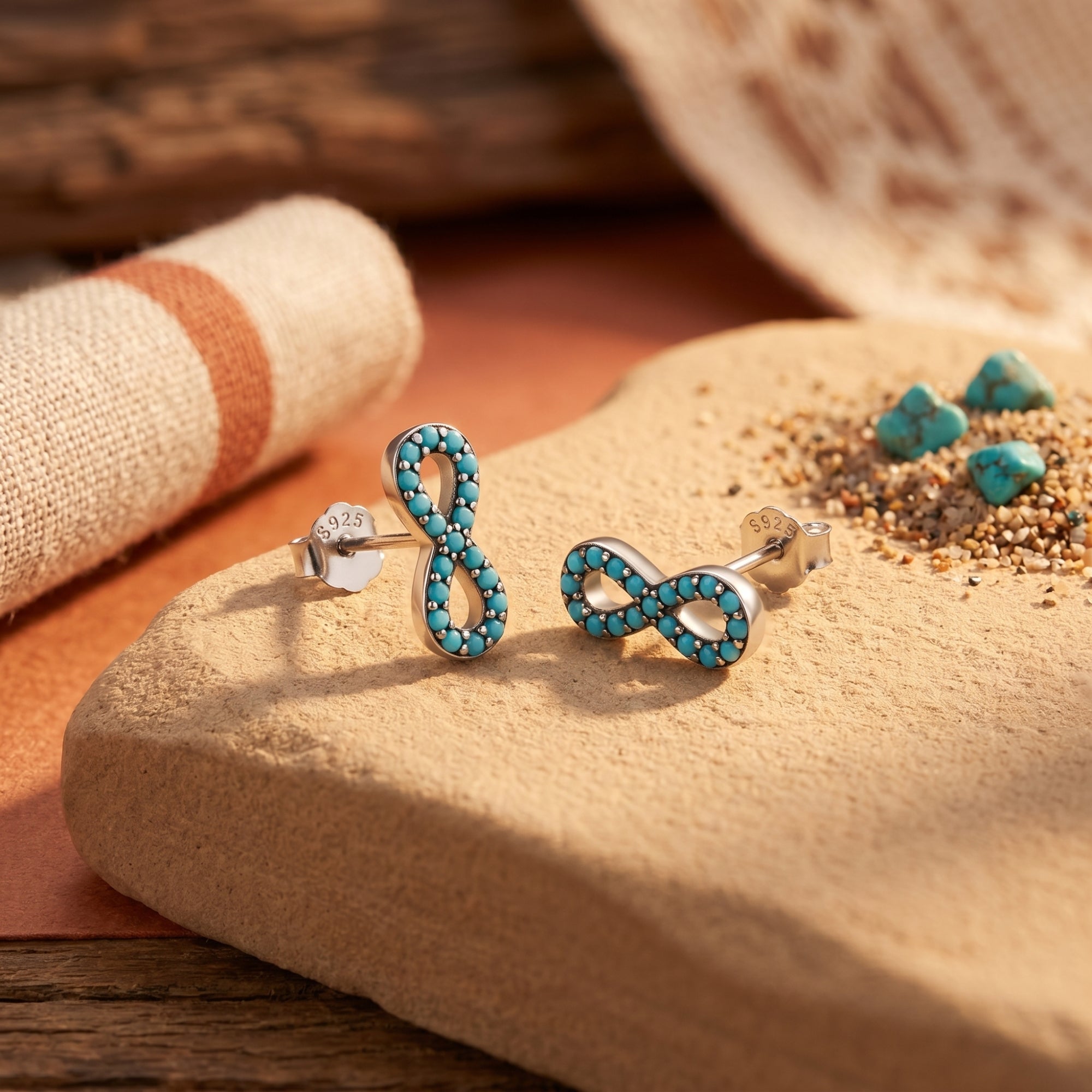 Silver infinity stud earrings with round turquoise stones on a desert-themed stone surface.