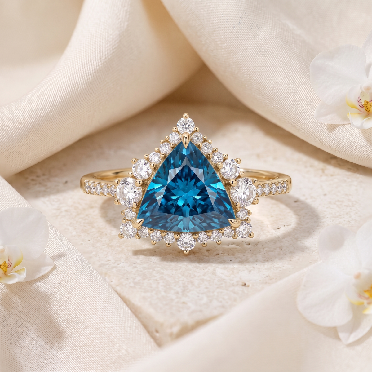 Trillion-cut blue moissanite halo engagement ring in yellow gold displayed on textured stone with draped fabric.
