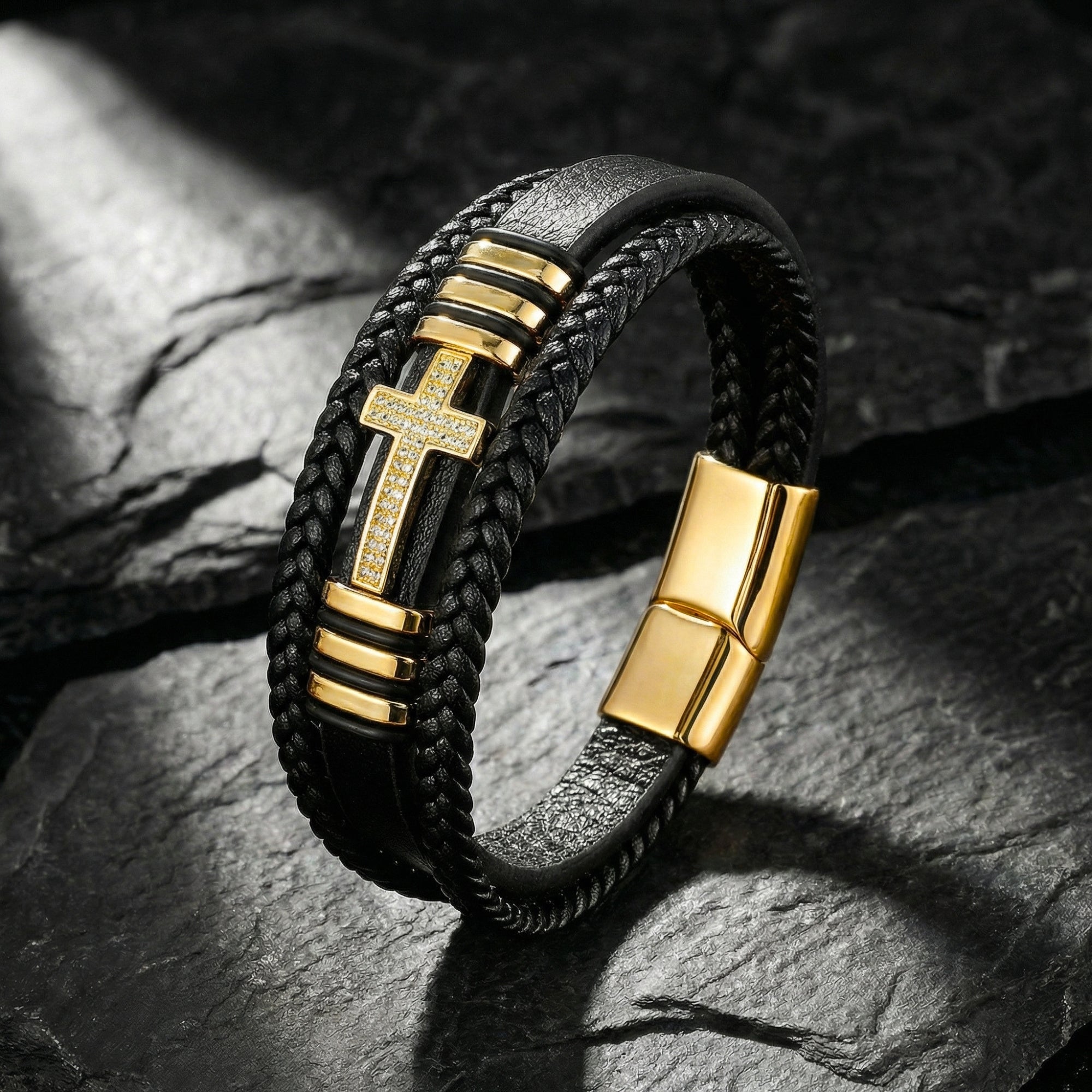 Gold-finished stainless steel cross bracelet with pavé stones on black braided leather, set on dark stone.