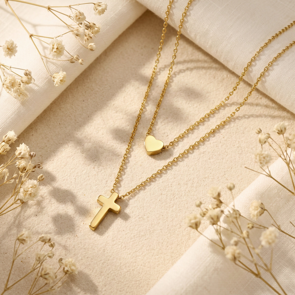 Flat lay of the layered gold-tone necklace with heart charm and cross pendant on sand.