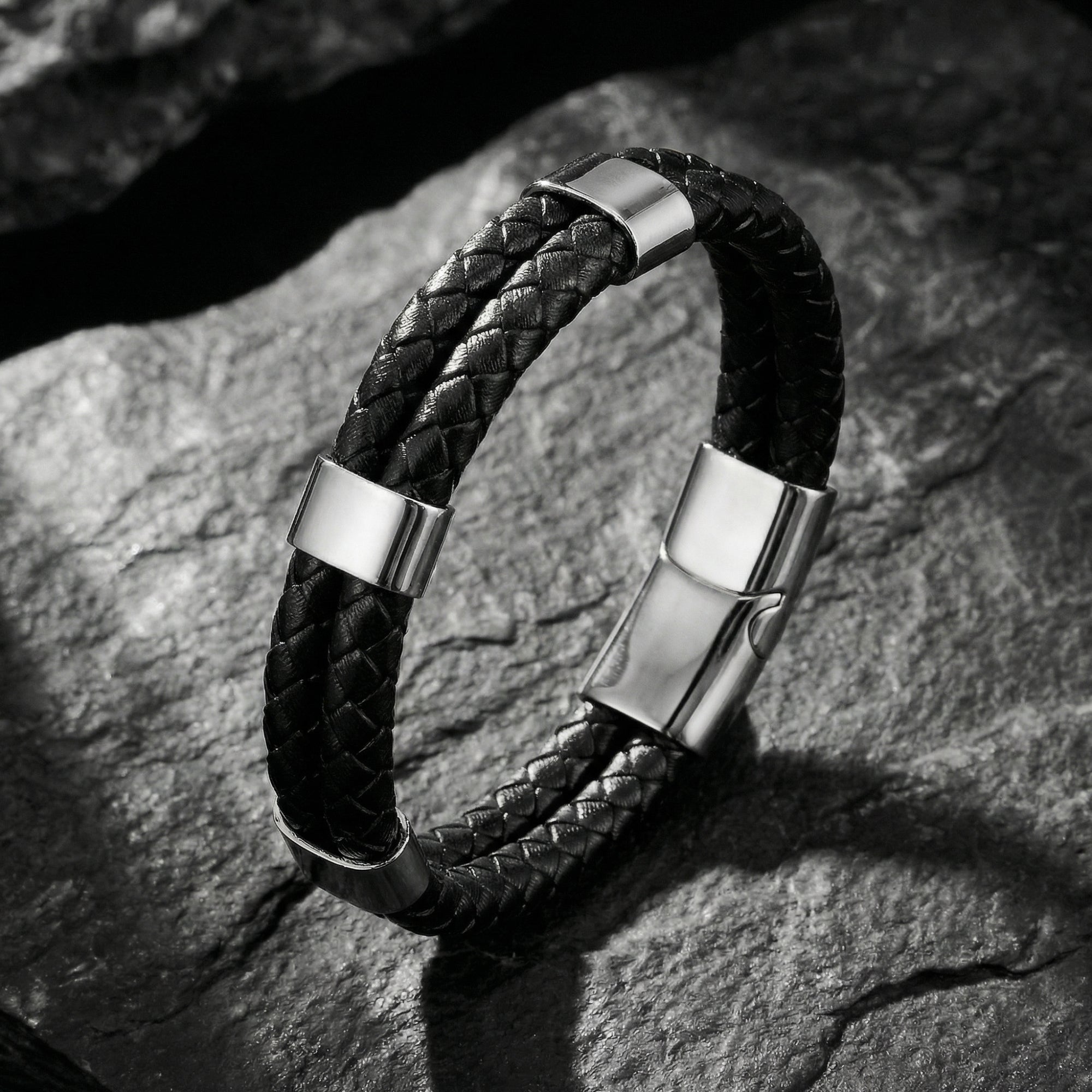 Black double-braided leather Sentinel bracelet with silver stainless steel hardware on a dark rock background.