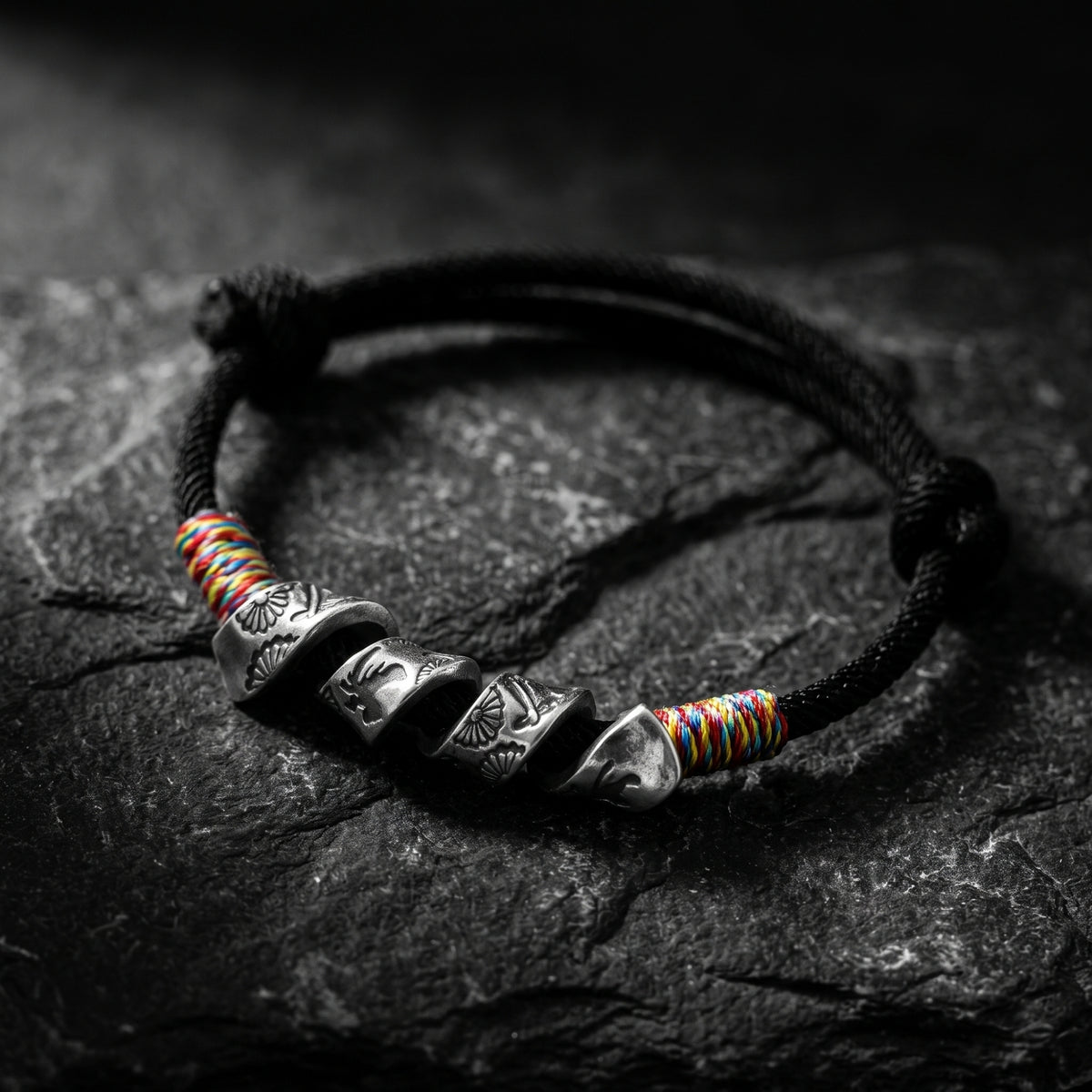 Black cord bracelet with multiple engraved silver curved sections and multicolored thread accents on dark stone.
