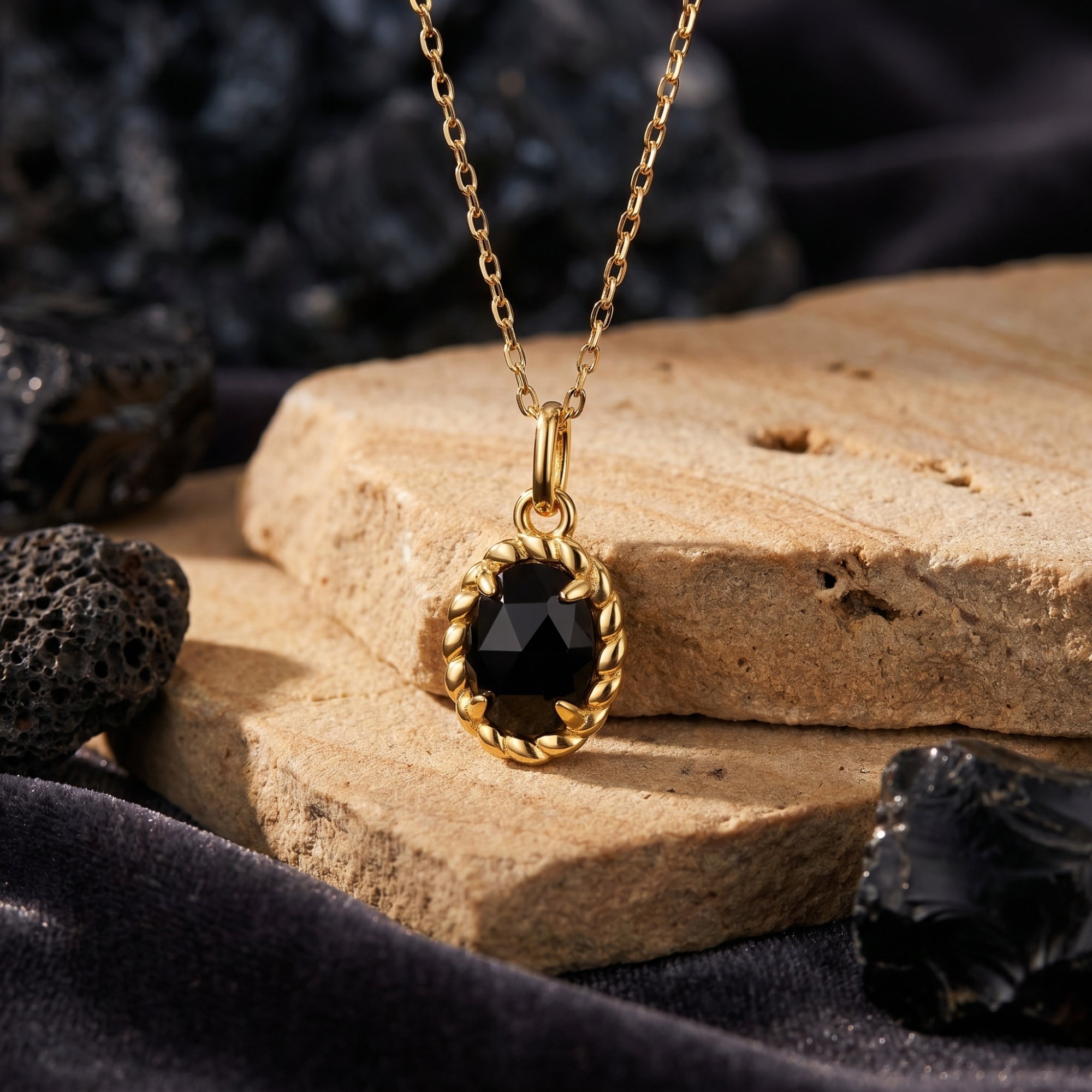 Oval-cut black onyx pendant in a yellow gold twisted setting, styled on a natural stone with dark volcanic rocks.
