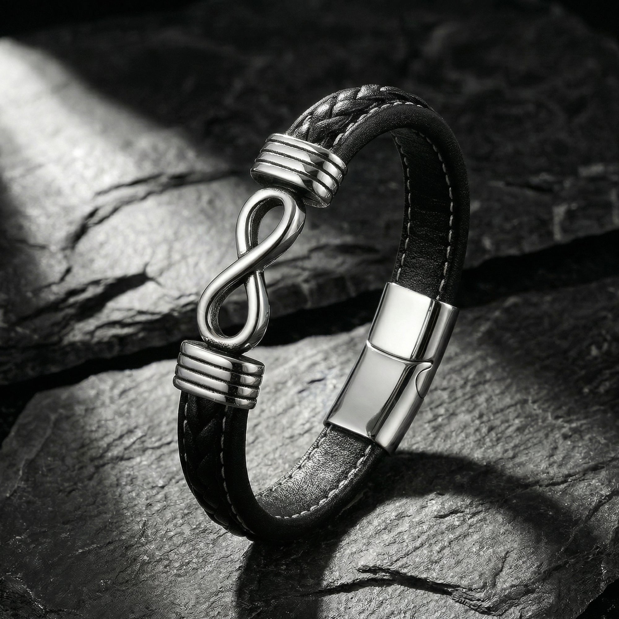 Men's black braided leather bracelet with a silver stainless steel infinity clasp, angled view on stone.