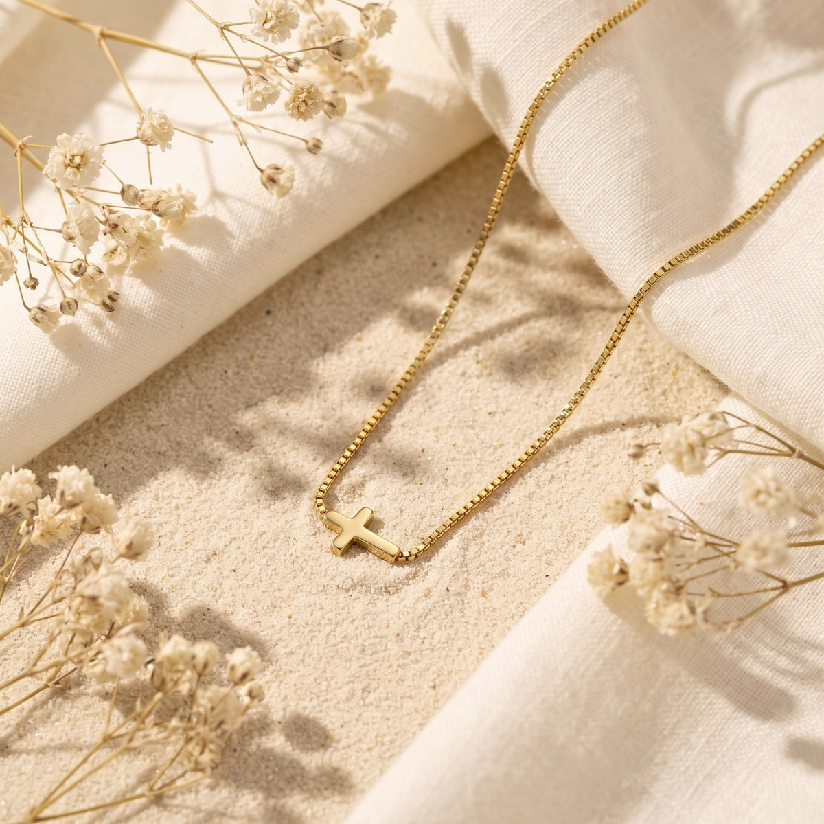 Gold-tone horizontal cross necklace resting on beige sand, surrounded by cream fabric and white dried flowers.