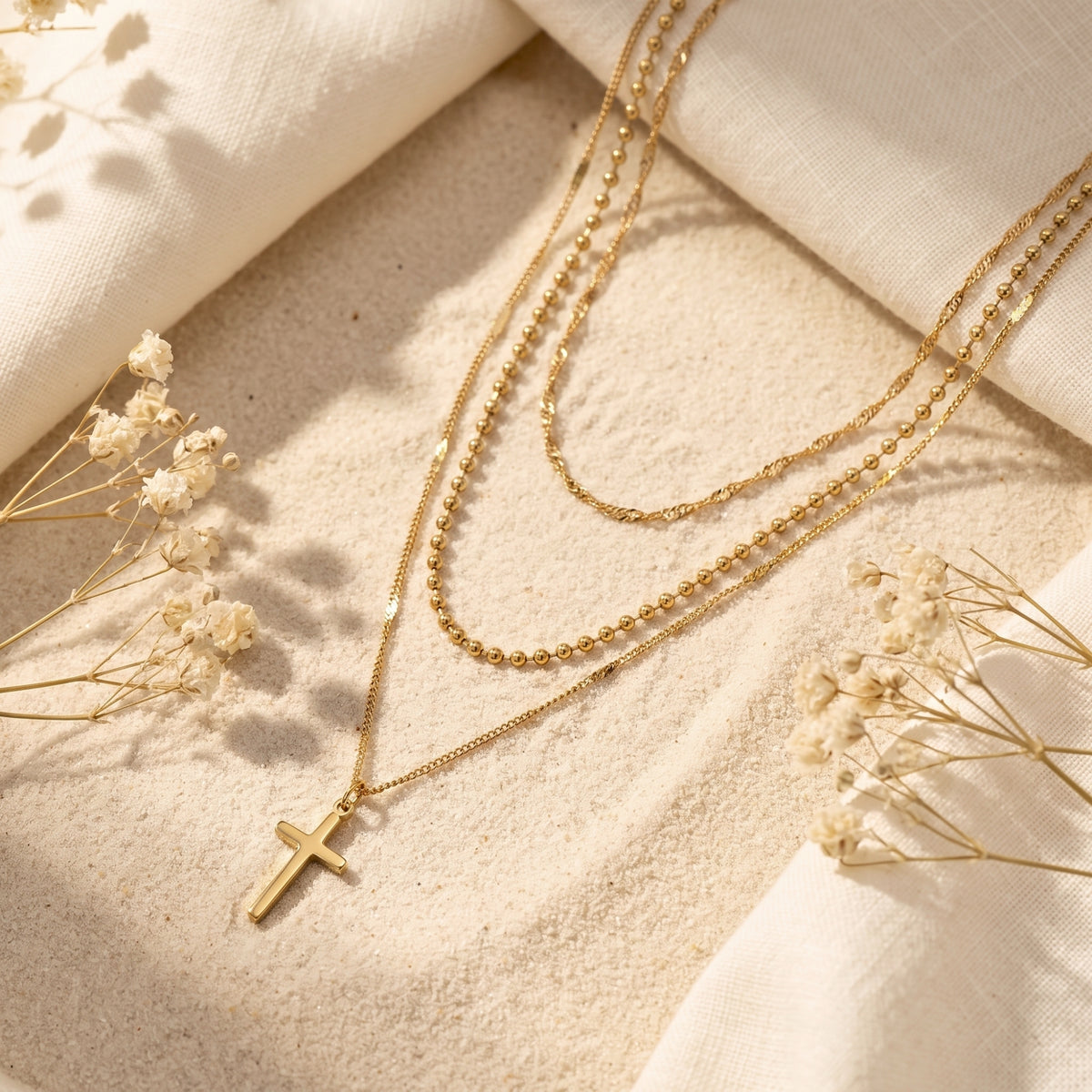 Gold-tone three-strand layered necklace with cross pendant lying flat on sand beside white fabric and dried flowers.