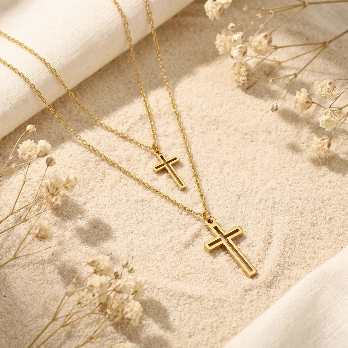 Gold-tone layered double cross necklace laid diagonally on pale sand with white fabric and dried flowers.