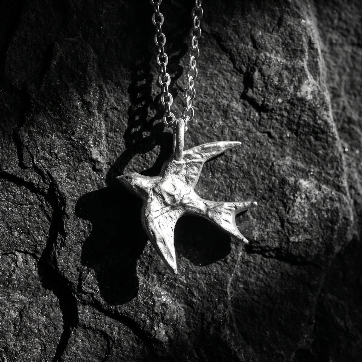 Textured silver swallow pendant on a link chain against a dark, rough stone surface.