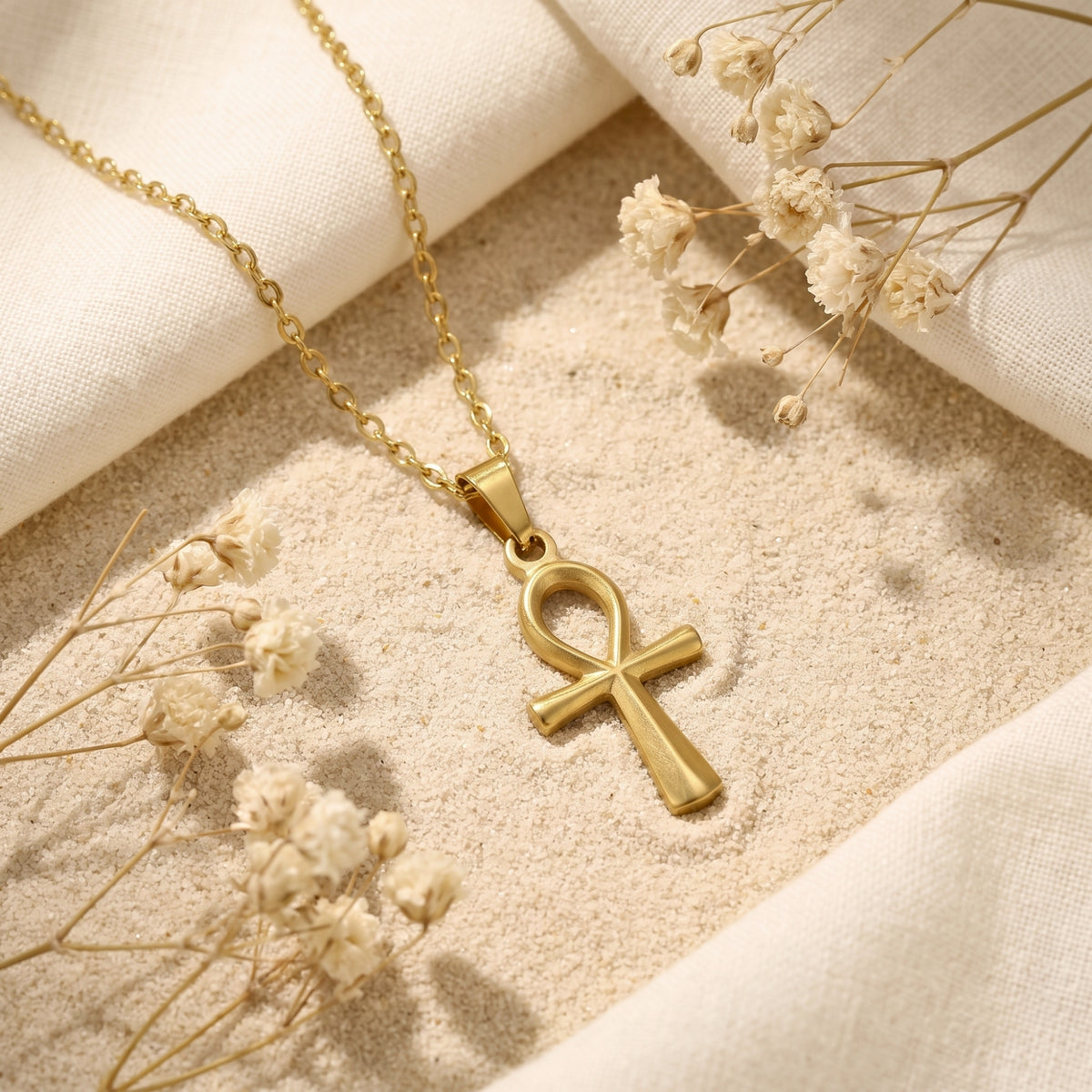 Gold-tone Ankh cross pendant necklace resting on sand with dried flowers and fabric.