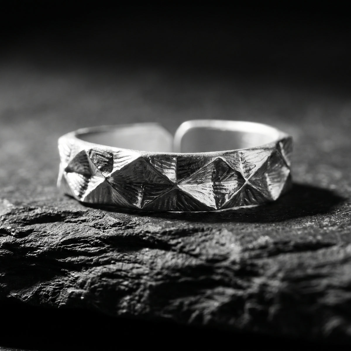 Silver faceted pyramid open-band ring resting on dark textured rock in a close-up studio view.