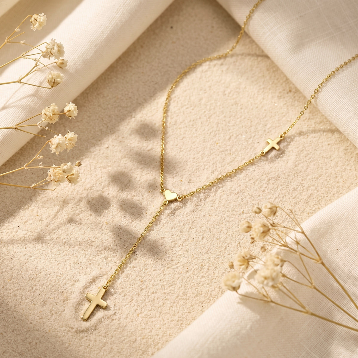 Flat lay view of the gold-tone lariat necklace with heart centerpiece, drop cross, and side cross on a sandy surface.