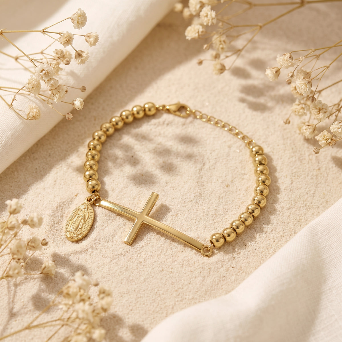 Gold-tone beaded bracelet with a horizontal cross bar and Virgin Mary charm, shown flat on sand.