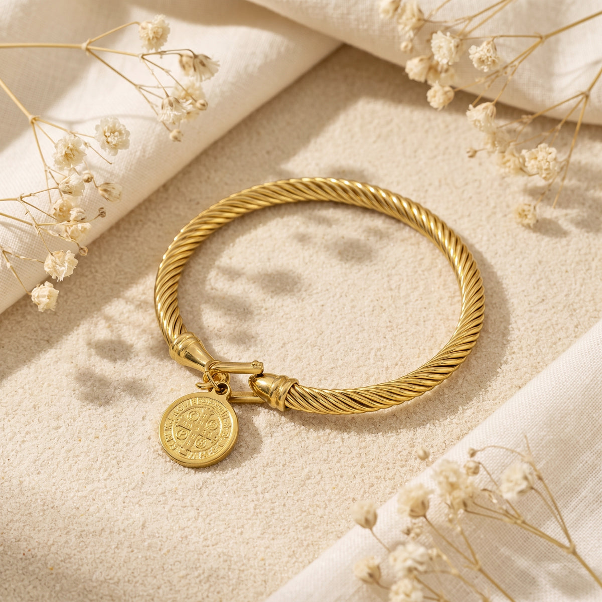 Gold-tone twisted cable bracelet with a circular medal charm, shown flat on sand surrounded by white linen and dried flowers.